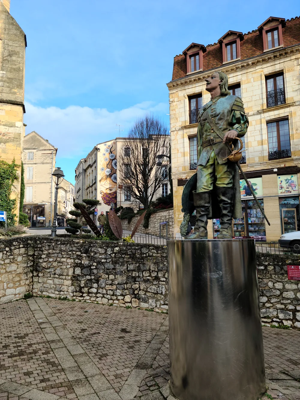 Bronze statue of Cyrano de Bergerac on a stone plinth in Place Pélissière, medieval stone buildings rising behind it under blue sky