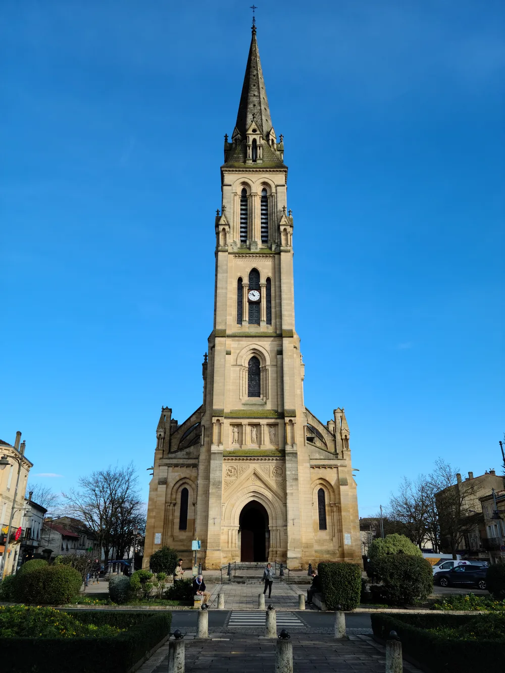 Tall Gothic spire of Notre-Dame church in Bergerac rising above its ornate stone facade against a brilliant blue sky, steps and shrubs in the foreground