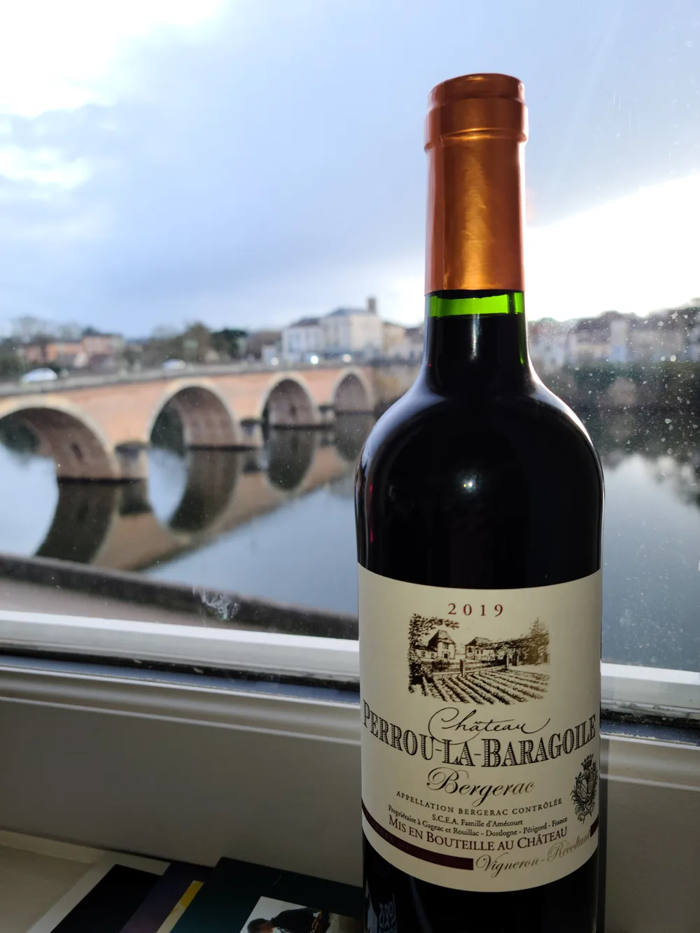 Bottle of 2019 Château Perrou-la-Baragoile Bergerac red wine on a windowsill, the Pont Vieux arches and Dordogne River visible in the background