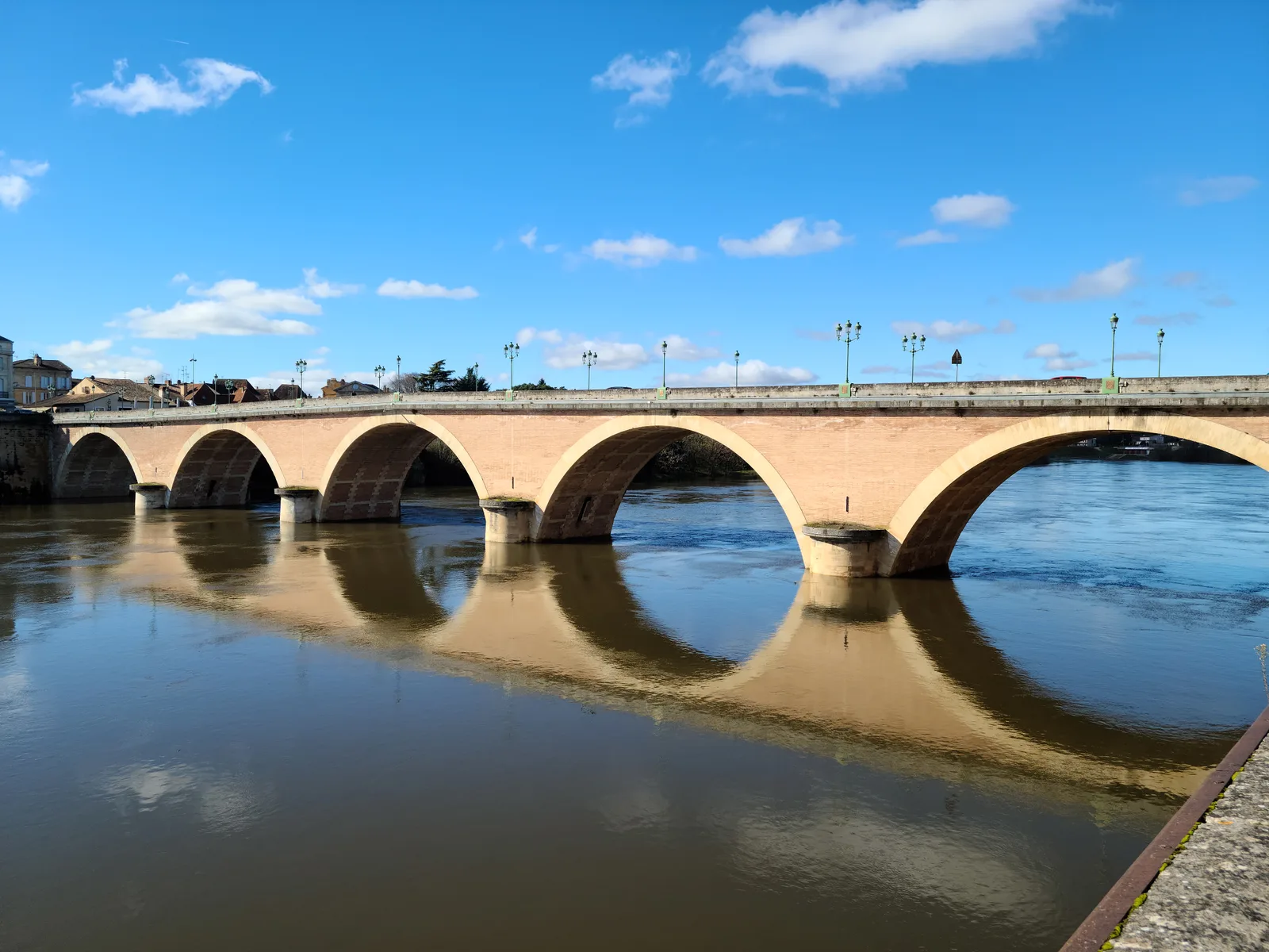 The stone arches of Bergerac's Pont Vieux perfectly reflected in the still Dordogne River under a blue sky with scattered white clouds