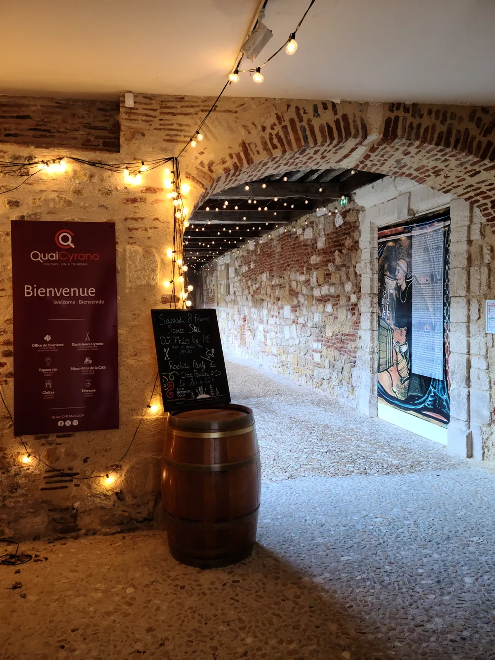 Stone arched corridor at the Quai Cyrano wine cave in Bergerac, strung with warm fairy lights, a wine barrel at the entrance and a Bienvenue welcome sign