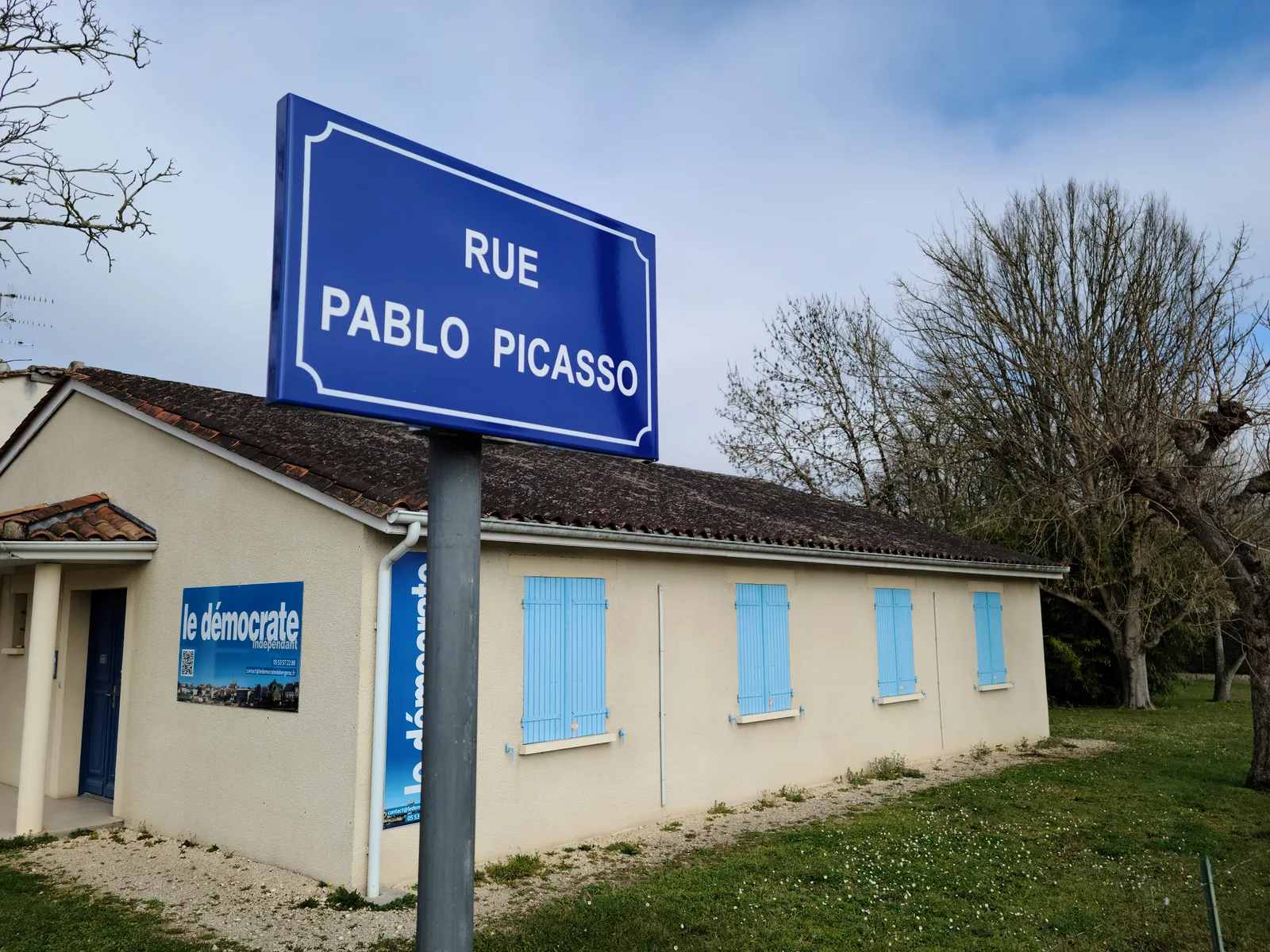 Blue French street sign for Rue Pablo Picasso on a post in Bergerac, with a cream building with blue shutters in the background