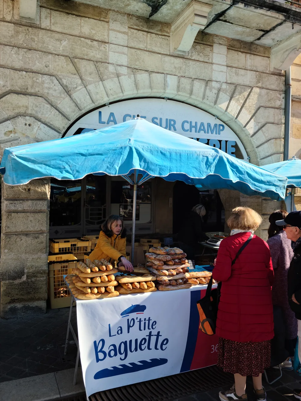 La P'tite Baguette stall at Bergerac's Saturday market with rows of fresh baguettes under a blue canopy, a vendor serving customers outside a stone building