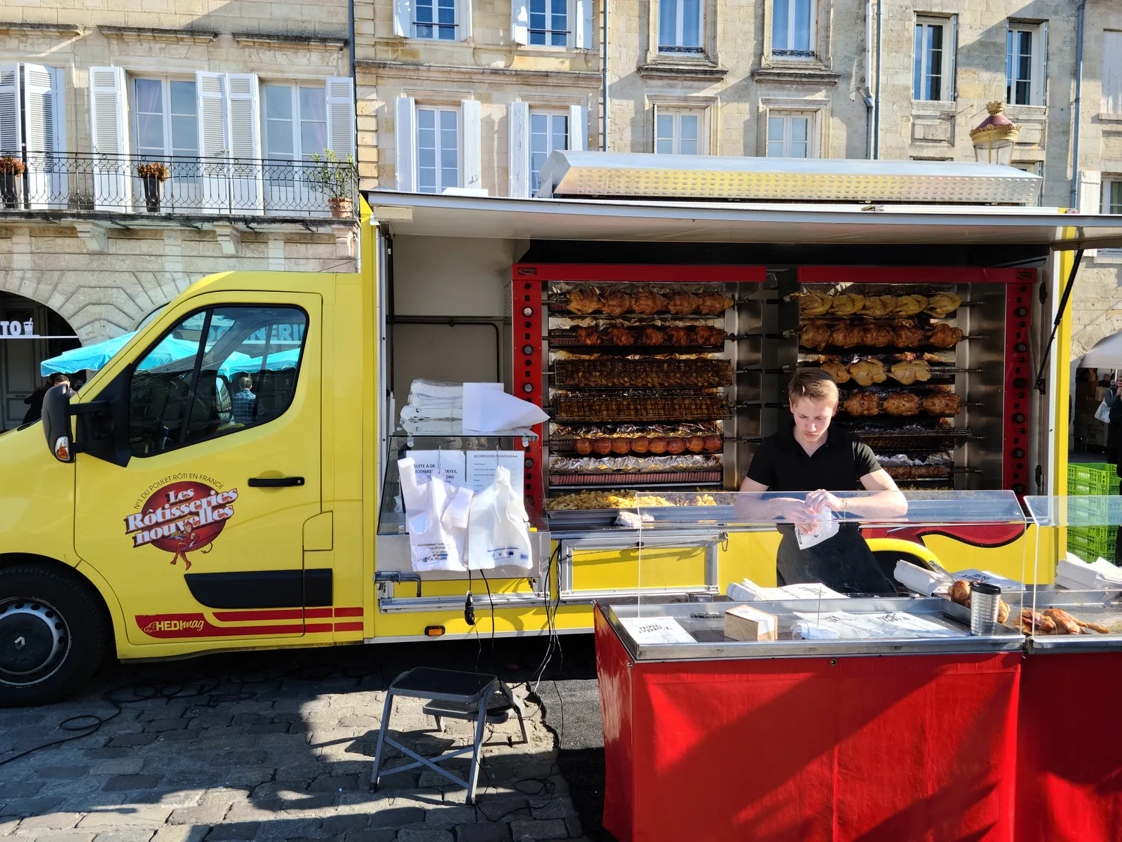 A yellow rotisserie truck at Bergerac's Saturday market with rows of chickens rotating on spits, a vendor serving customers on a sunny day