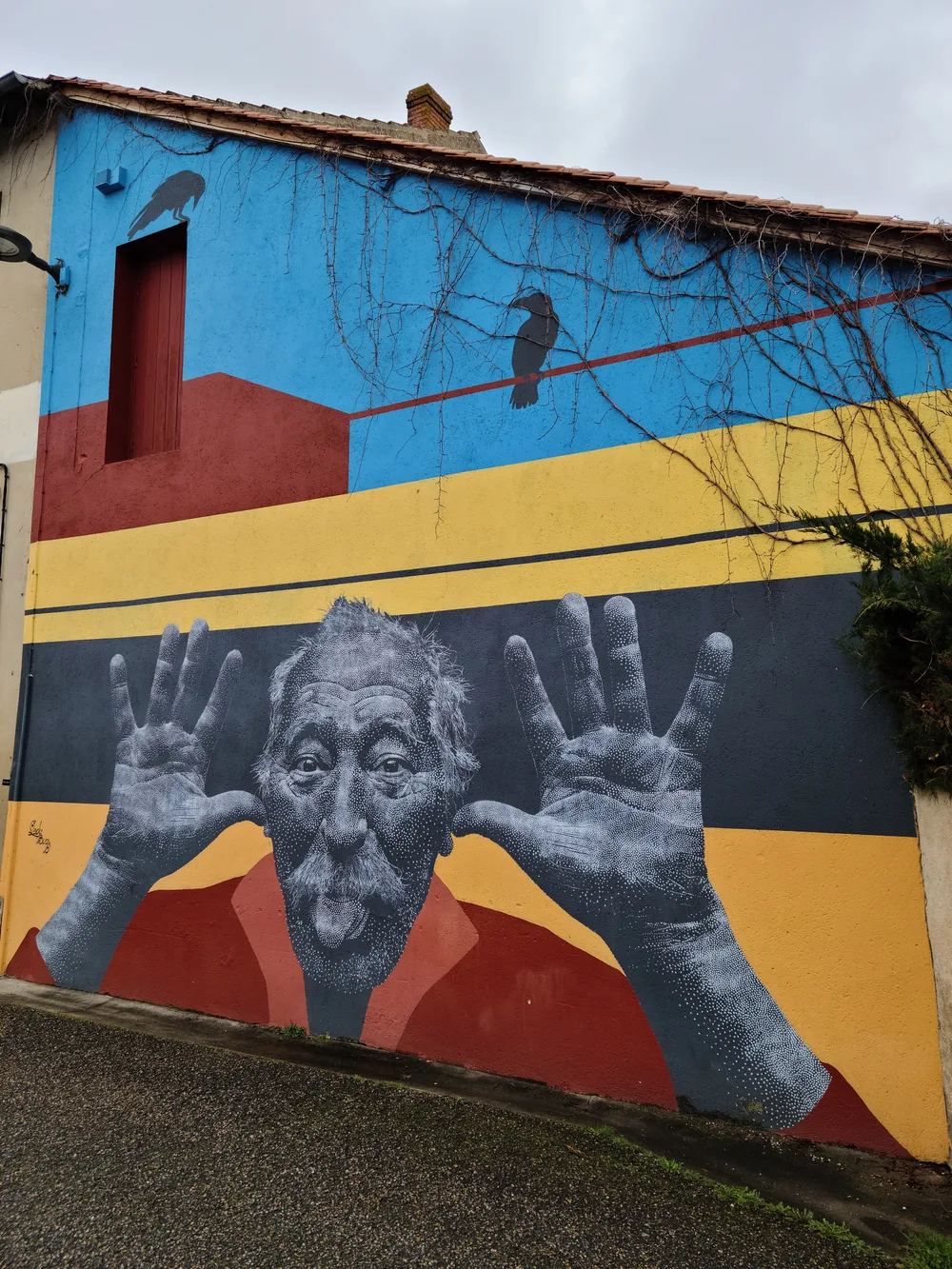 Street art mural on a Bergerac building depicting an elderly man with large open hands against bold blocks of yellow, red, and blue, a bird perched on a wire above