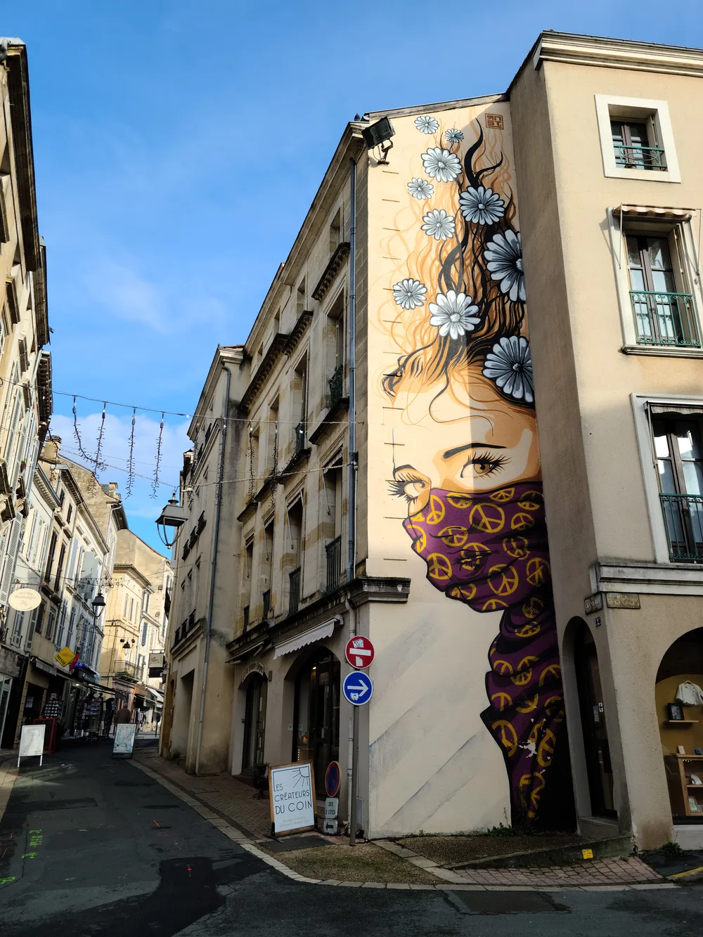 Large street art mural covering the corner of a Bergerac building depicting a woman's face with white daisy flowers in her hair and a patterned scarf