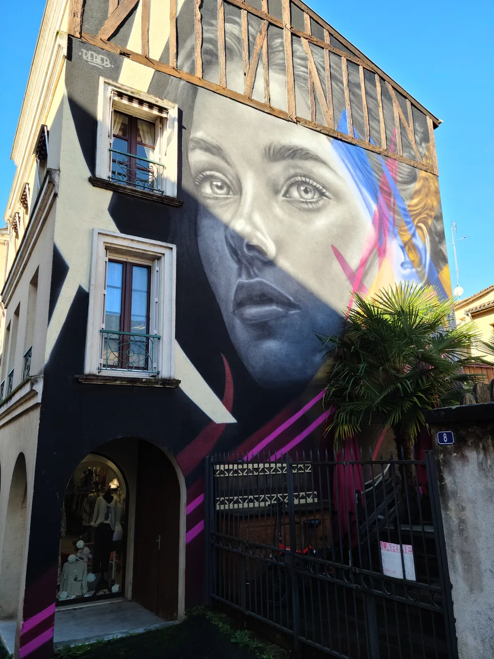 Photorealistic street art mural covering a tall building in Bergerac's old town depicting a woman's face with striking eyes, a shop at ground level below