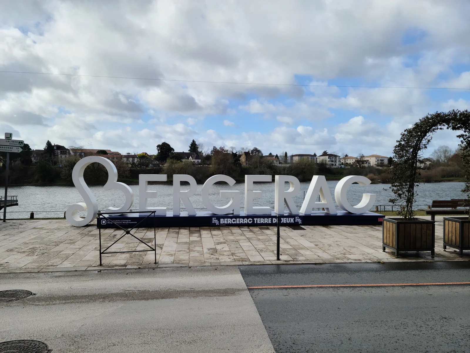Large white Bergerac letters installation on the Dordogne riverfront promenade, reading Bergerac Terre de Jeux under a cloudy sky