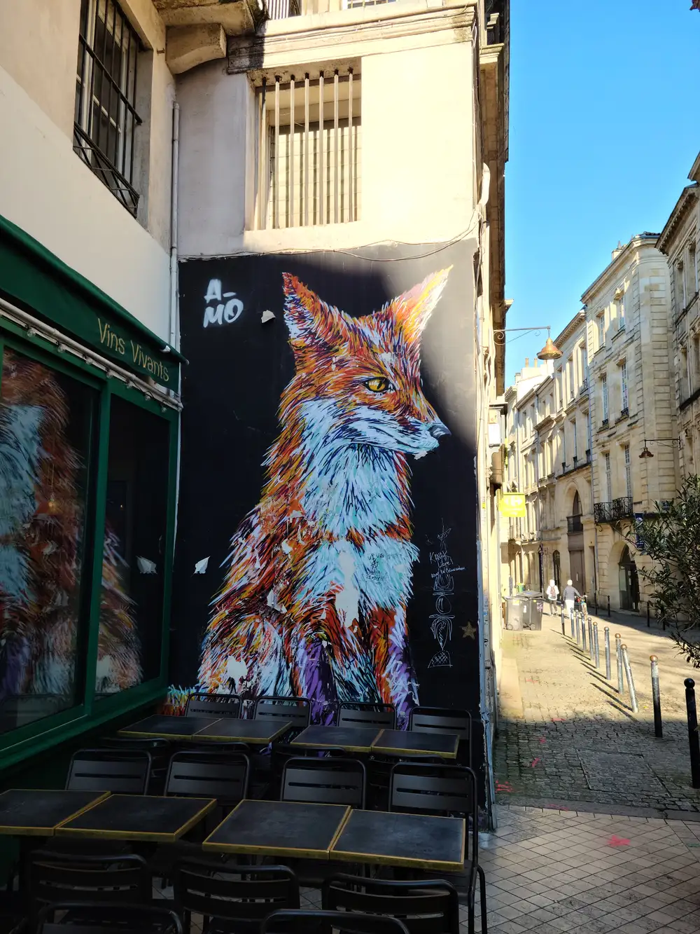Vibrant A-MO street art mural of a red fox towering above the Vins Vivants wine bar terrace on a sunny Bordeaux lane