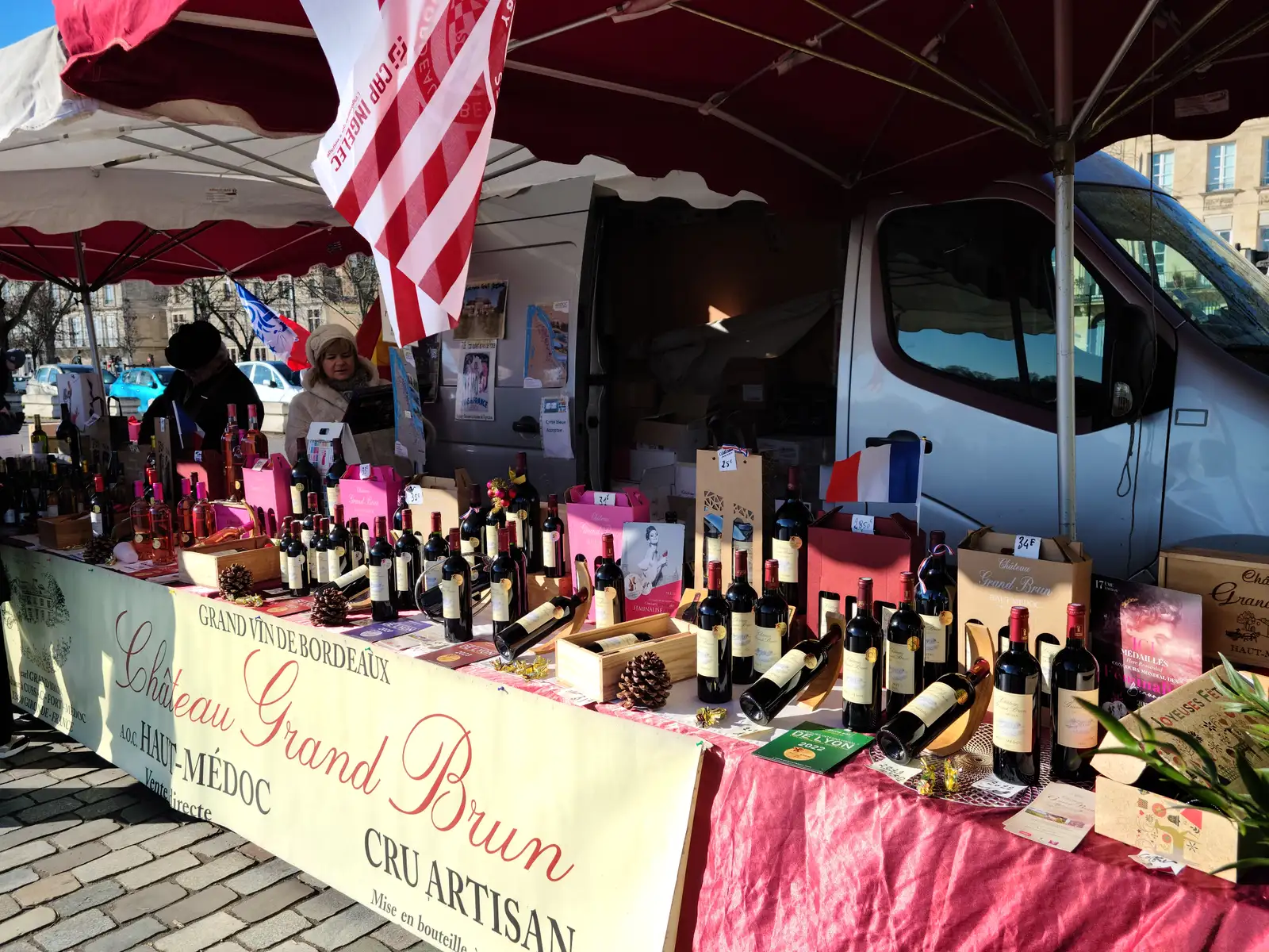 Château Grand Brun Haut-Médoc wine producer stall with red Bordeaux bottles and tricolor flags at an open-air Bordeaux market