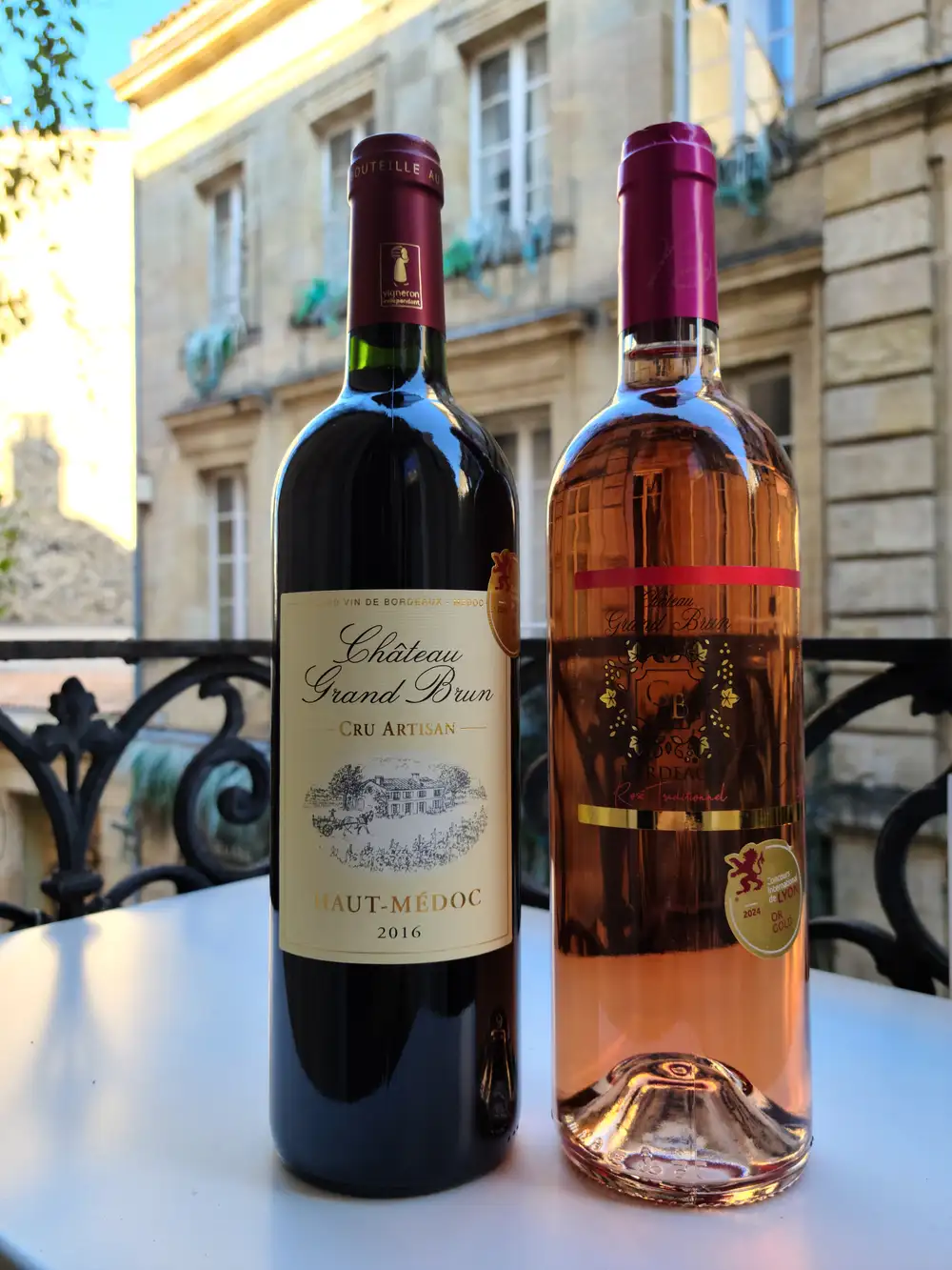 Bottles of Château Grand Brun Haut-Médoc red and rosé Bordeaux wines on a balcony table overlooking limestone facades