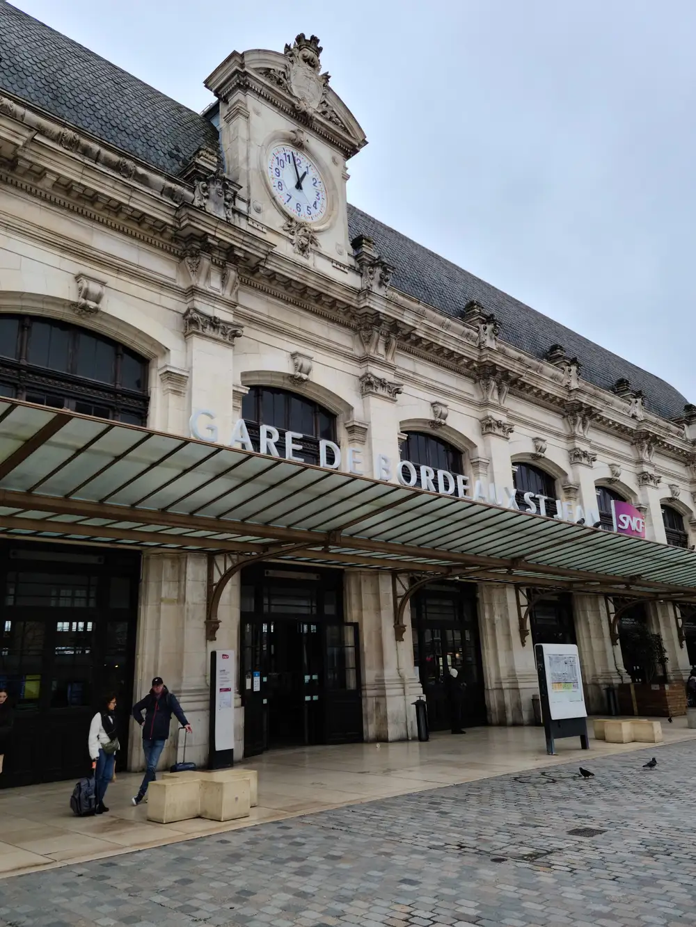 Travelers with luggage pass beneath the canopy of Gare de Bordeaux Saint-Jean below its ornate clock tower facade