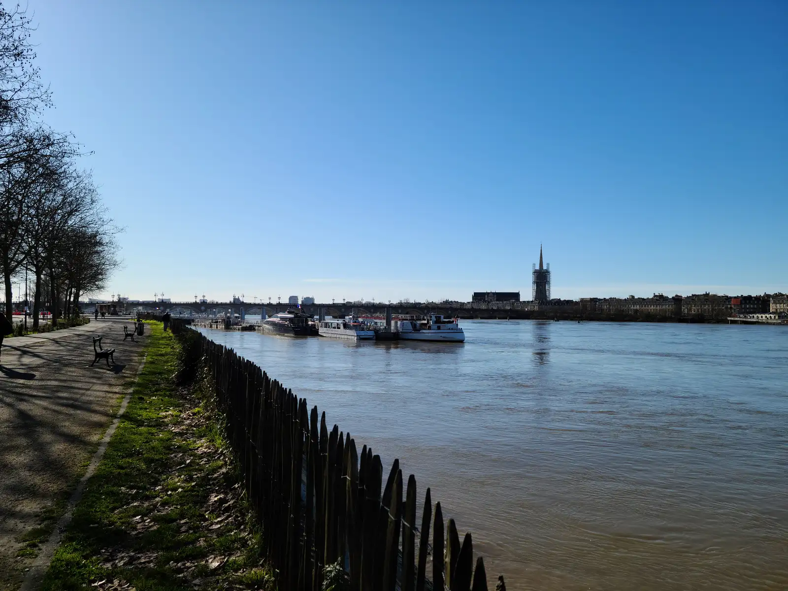 Riverside promenade with wooden fence and bench overlooking moored cruise boats and the Pont de Pierre on the Garonne in Bordeaux