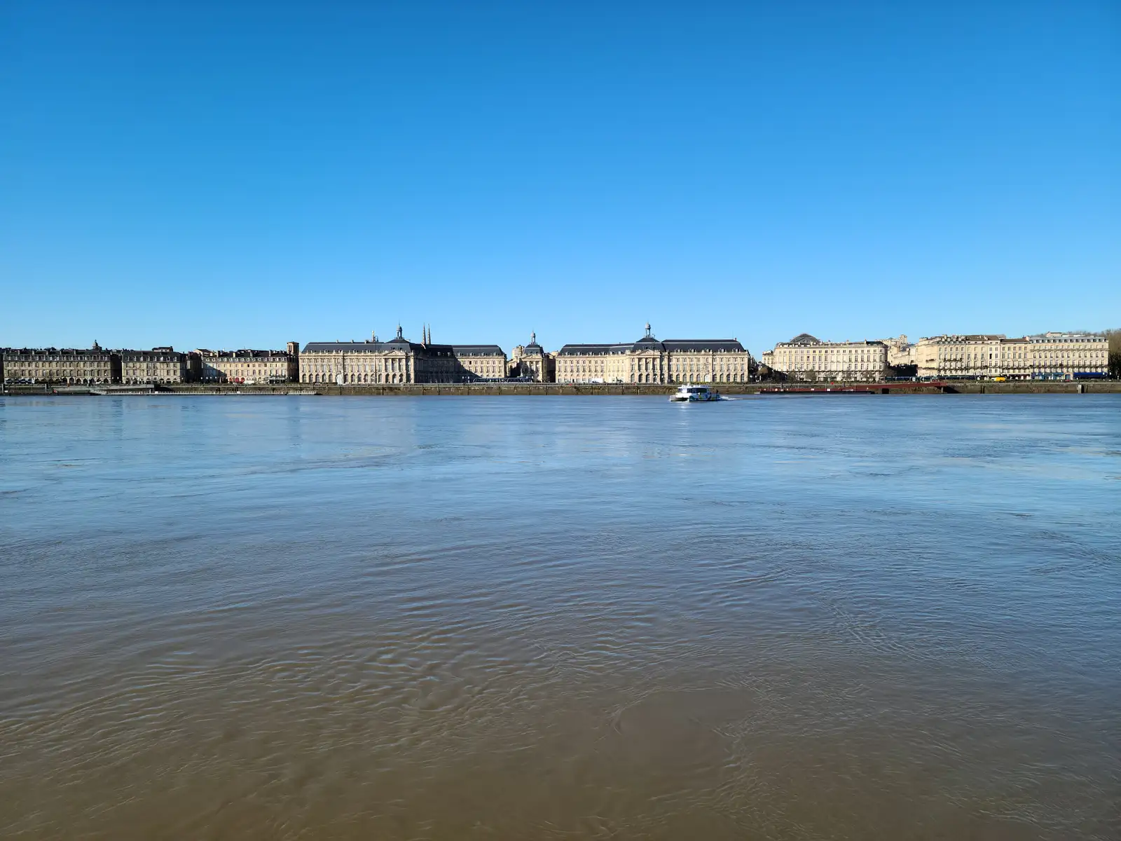 Wide view across the Garonne river toward the neoclassical Place de la Bourse skyline with a passing ferry in Bordeaux