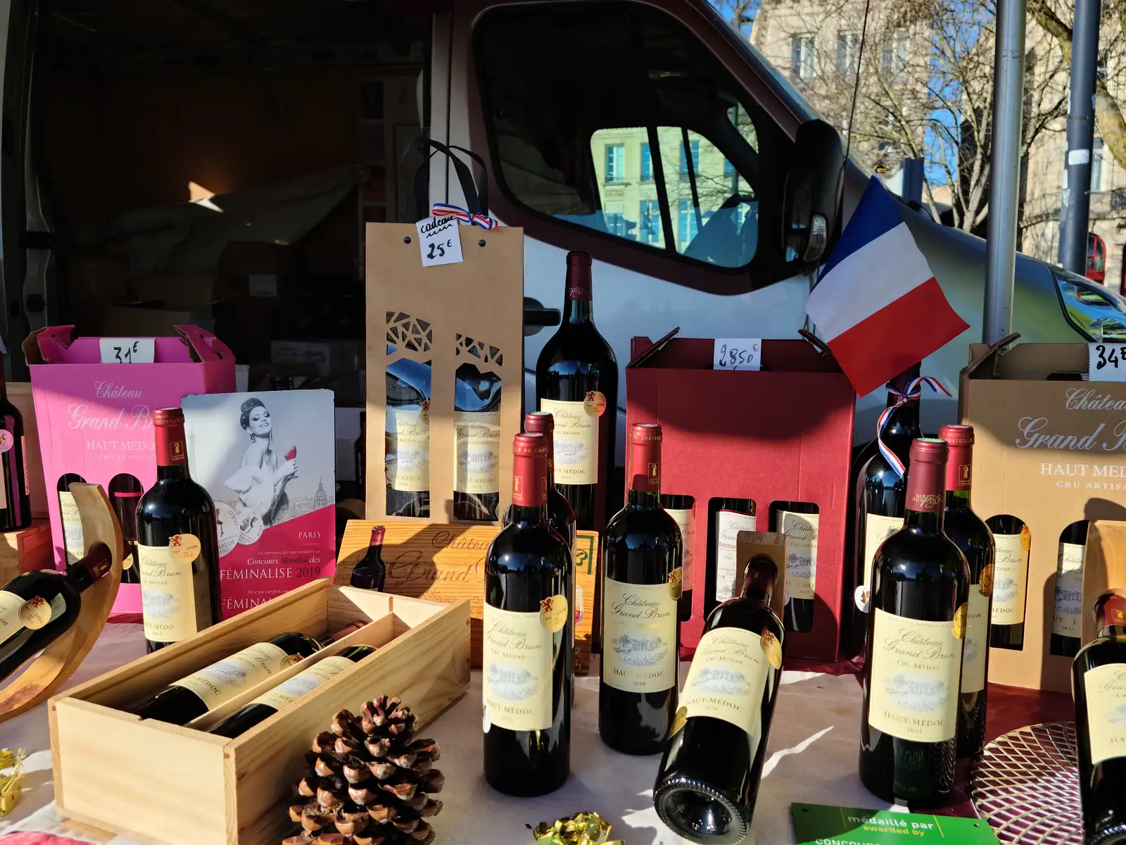 Red Bordeaux bottles from Château Grand Brun arranged with a French flag and wooden crates at a Haut-Médoc producer's market stall