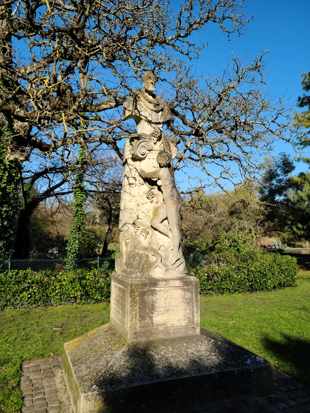 Weathered limestone statue of Bacchus with grape vine carvings on a pedestal beneath bare oak branches in Jardin Public, Bordeaux