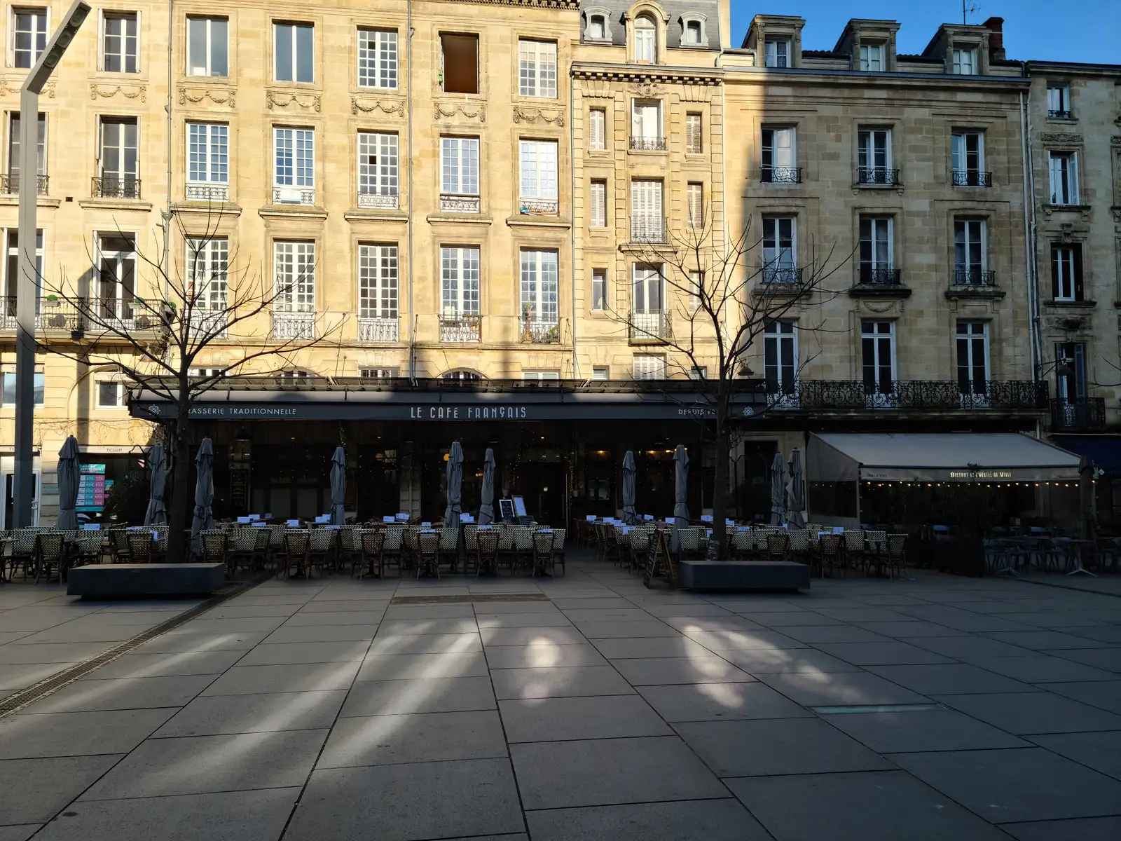 Le Café Français brasserie terrace with closed parasols and bistro chairs facing an empty sunlit square in Bordeaux