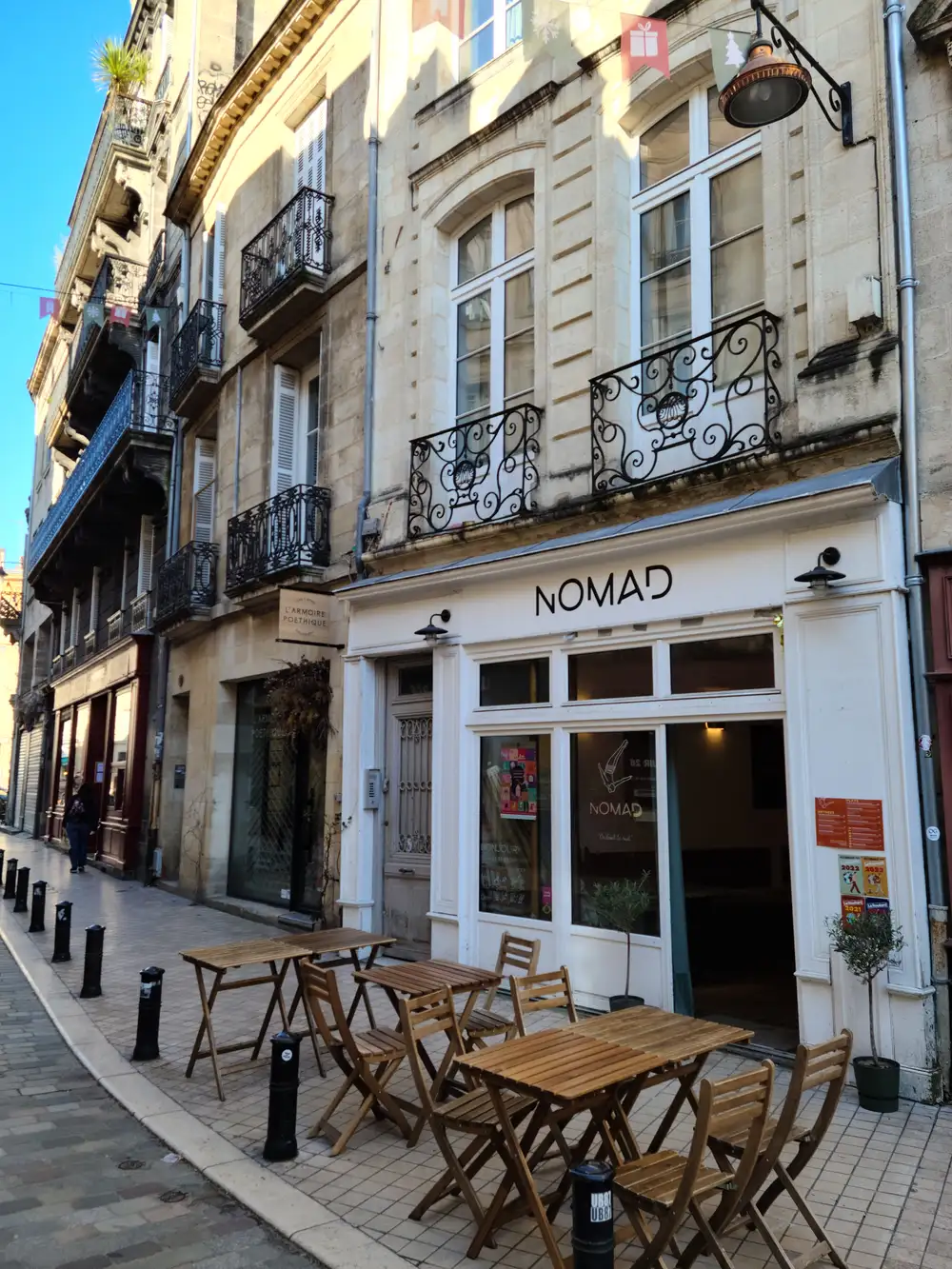 Nomad cafe terrace with wooden bistro tables beneath ornate wrought-iron balconies on a sunlit Bordeaux street