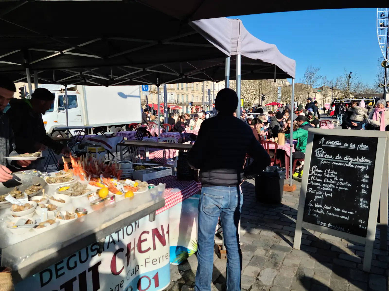 Customer in black jacket waits at a Cap-Ferret oyster tasting stall under a canopy at an open-air Bordeaux market