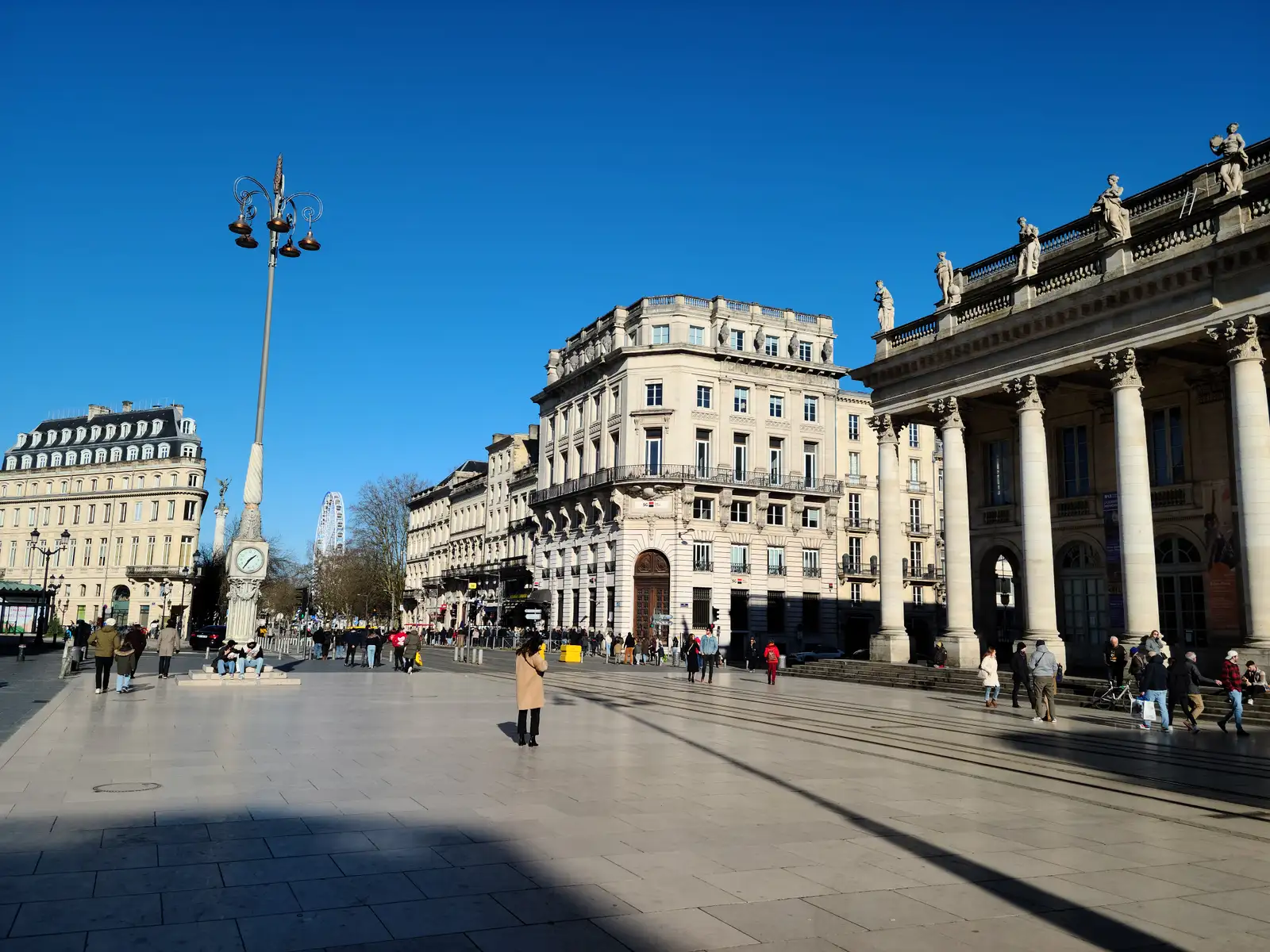 Ornate lamppost and clock tower on Place de la Comédie beside the Grand Théâtre colonnade under deep blue Bordeaux sky