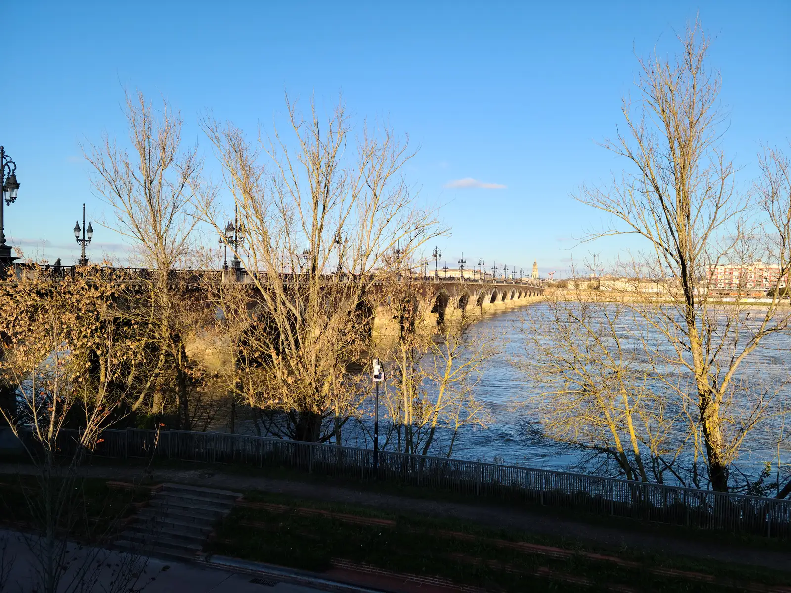 Pont de Pierre stone arches spanning the Garonne river framed by bare winter trees and lampposts under blue Bordeaux sky