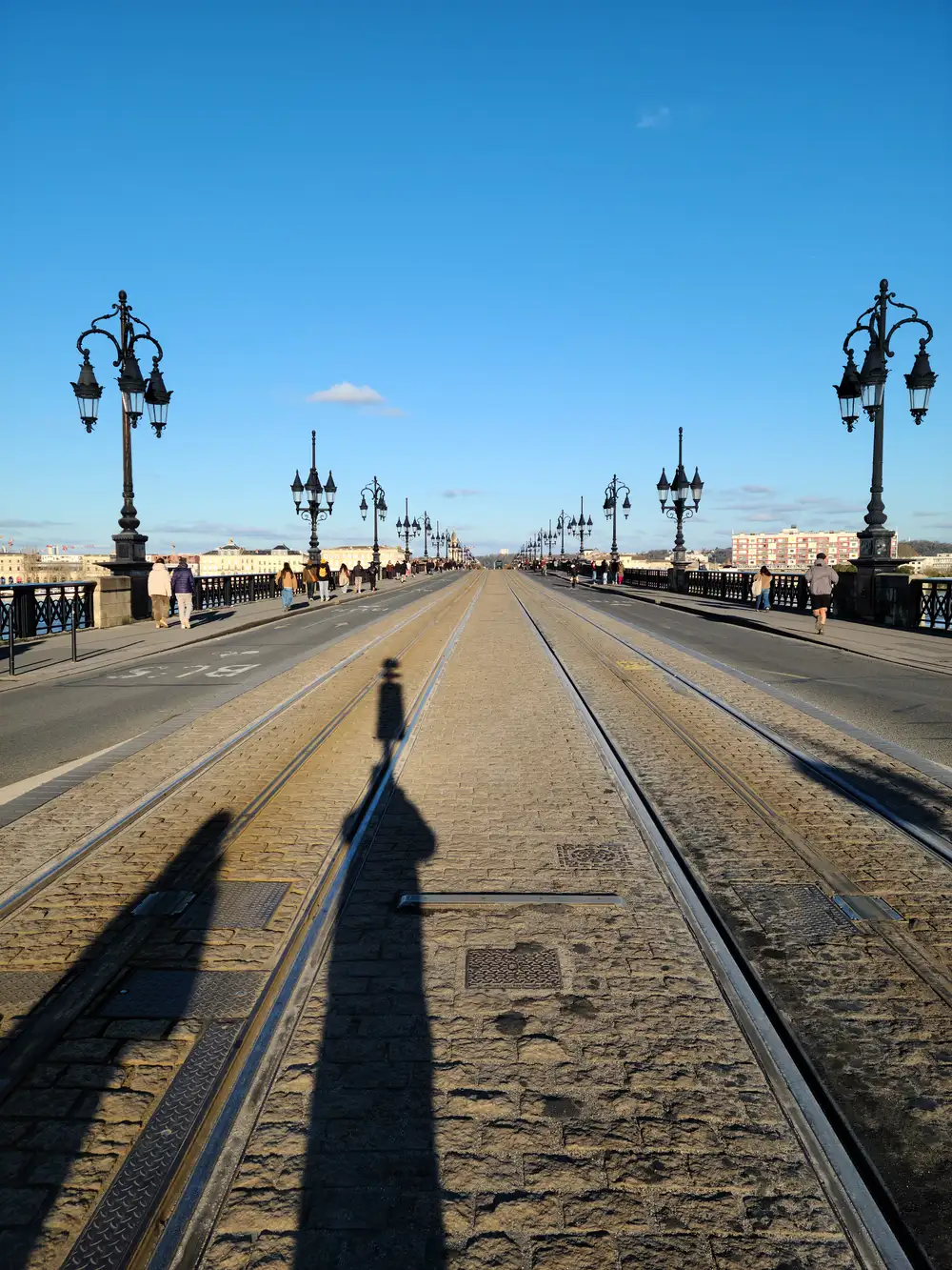 Photographer's shadow stretches along tram tracks on Pont de Pierre lined with ornate black lampposts in Bordeaux