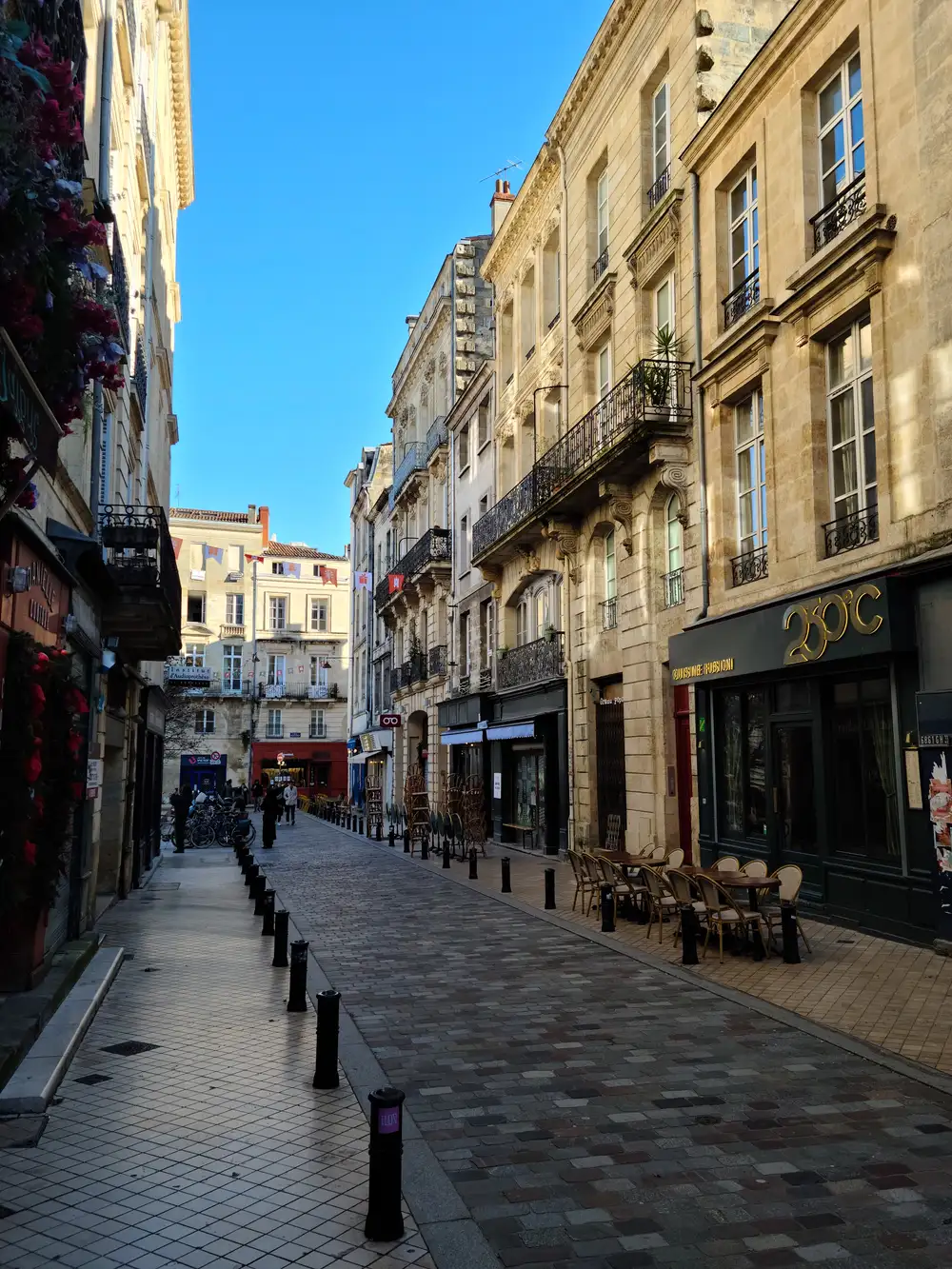 Sunlit pedestrian street lined with limestone buildings, wrought-iron balconies, and cafe terraces with bistro chairs in Bordeaux