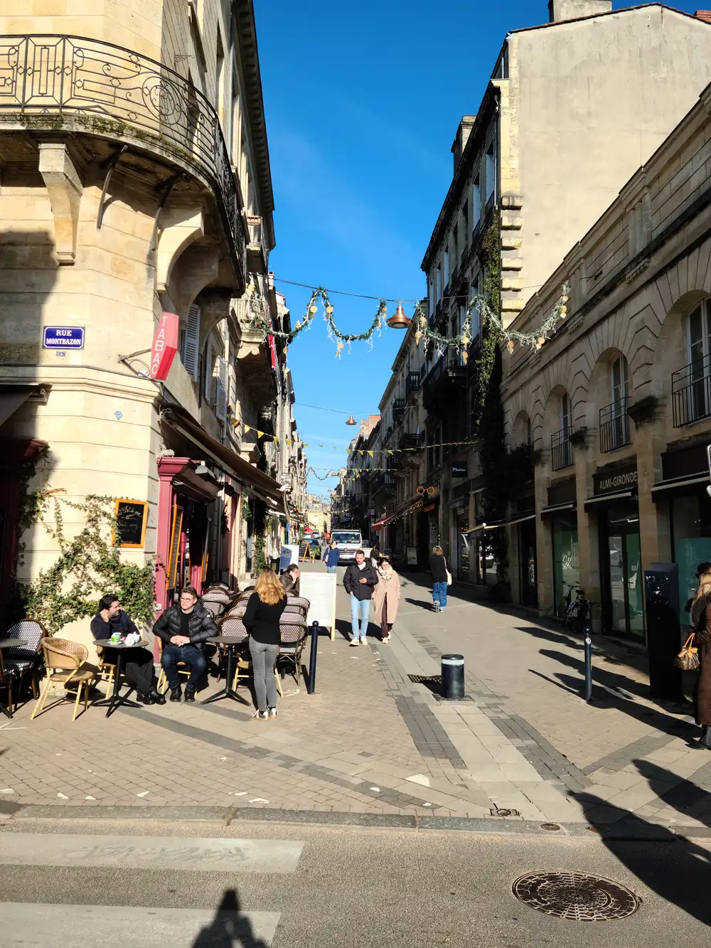 Sunlit corner of Rue Montrazon with a Tabac sign, cafe terrace, and festive garlands strung across a Bordeaux street