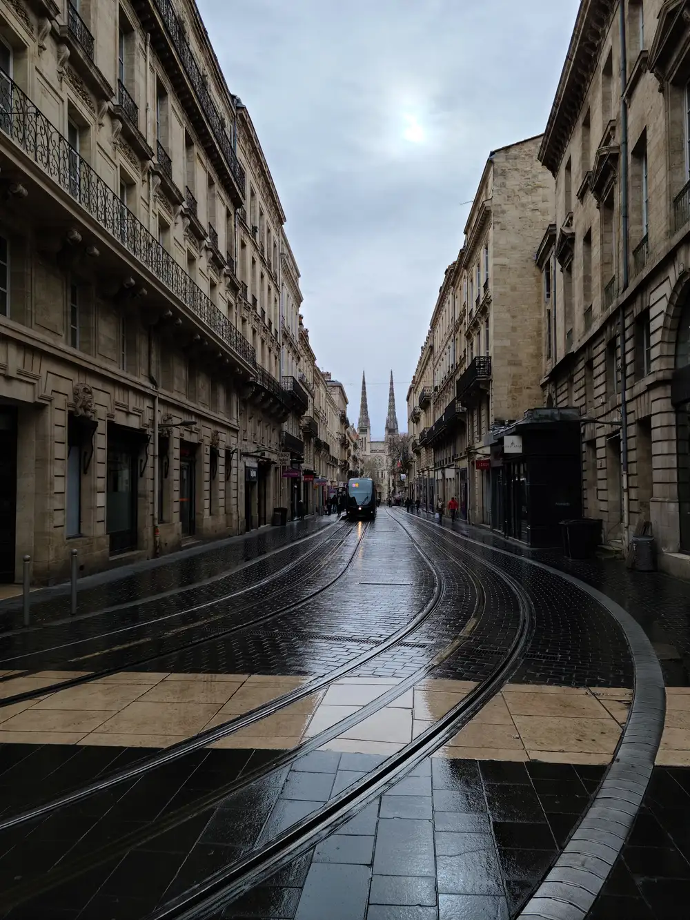Tram approaches on wet tracks along Rue Vital Carles framed by limestone facades and the cathedral spires beyond in rainy Bordeaux