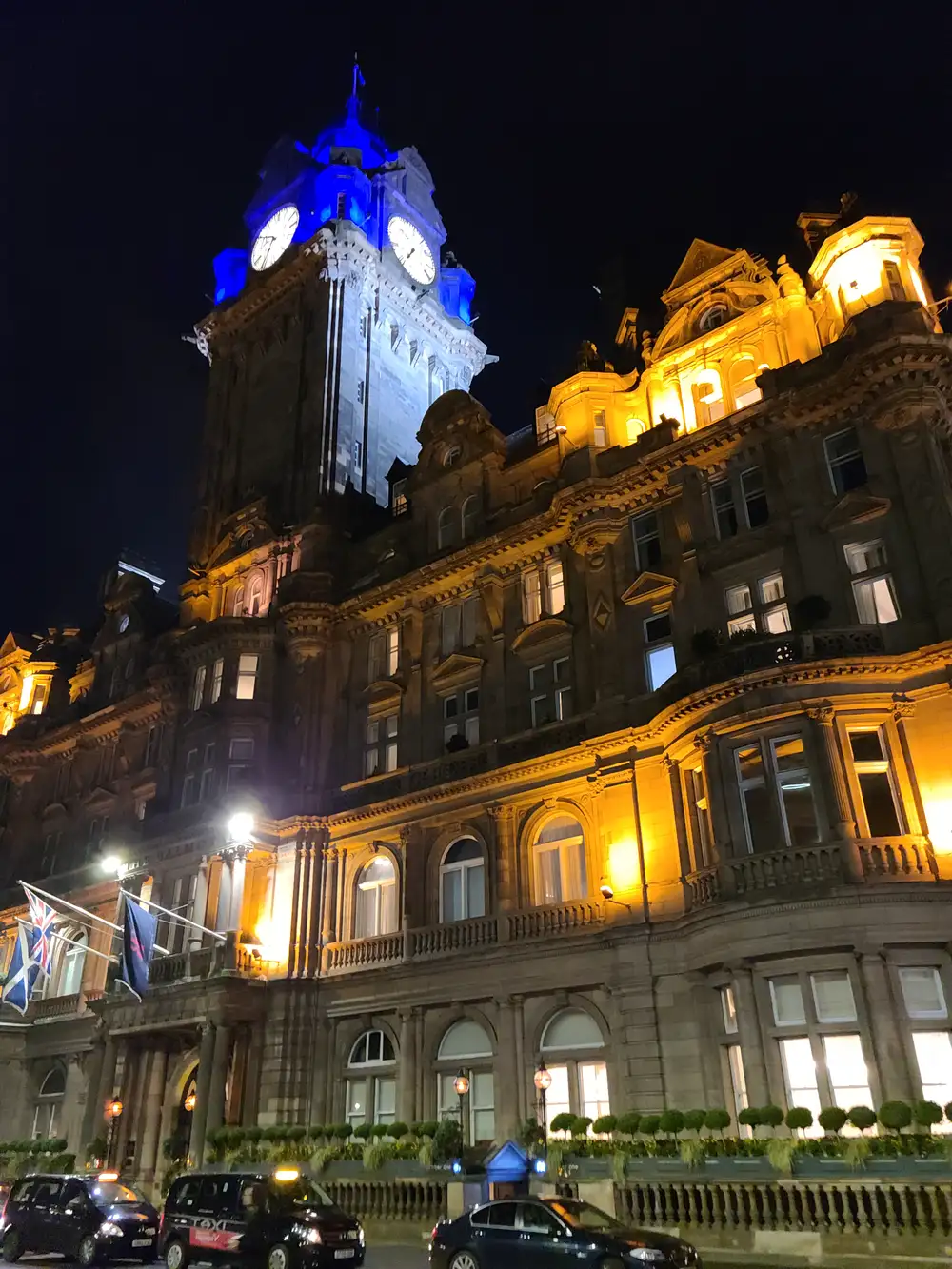 Balmoral Hotel's clock tower lit blue at night above the floodlit sandstone facade and Scottish flags on Princes Street Edinburgh