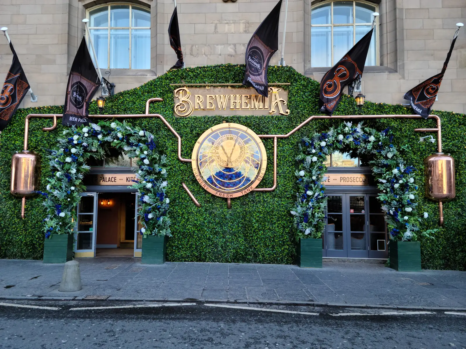 Brewhemia bar facade in Edinburgh with ivy-covered walls, ornate gold astronomical clock, copper piping and heraldic flags