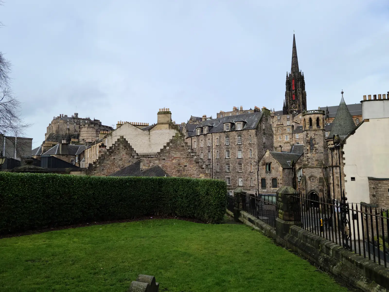 View from a grassy lawn in Edinburgh Old Town towards Edinburgh Castle on its crag and The Hub's gothic spire under gray skies