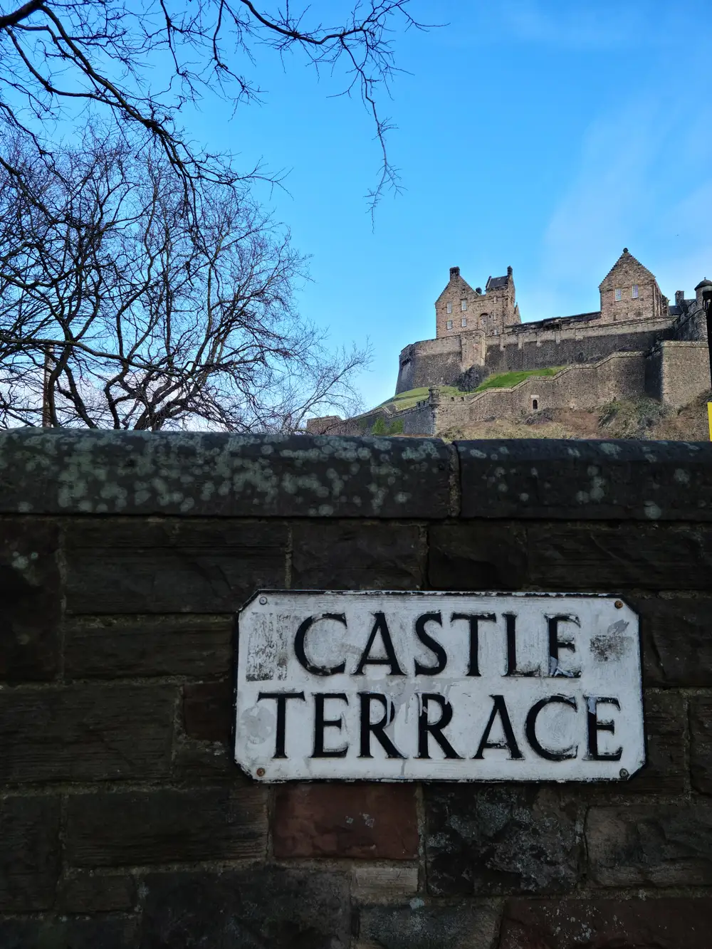 Vintage Castle Terrace street sign on a stone wall with Edinburgh Castle's ramparts rising above bare winter trees