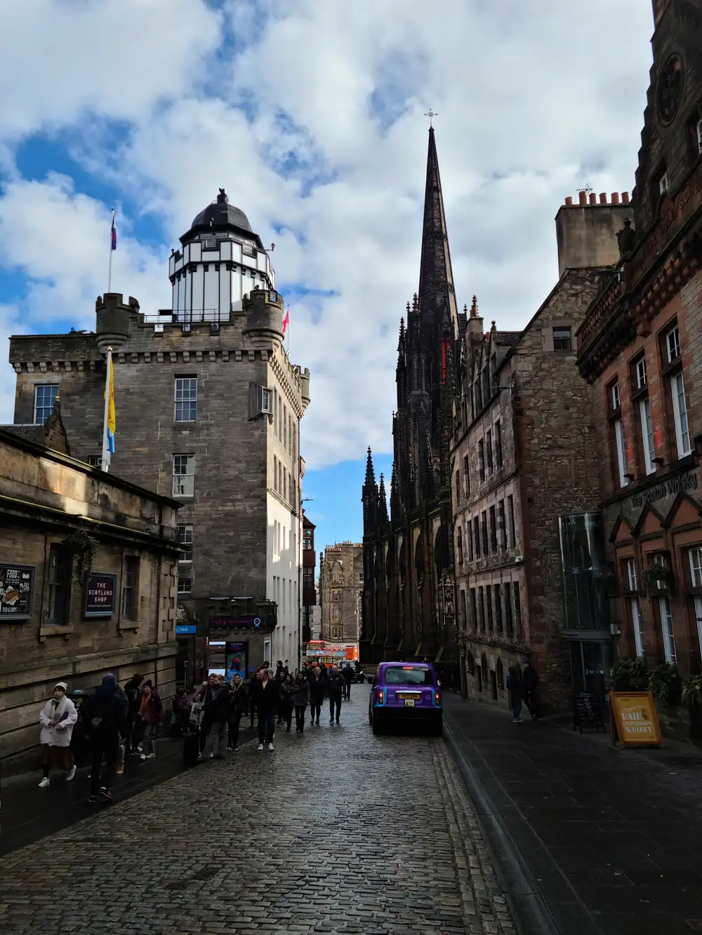 Purple taxi descending cobbled Castlehill on Edinburgh's Royal Mile with Camera Obscura tower and The Hub's gothic spire above