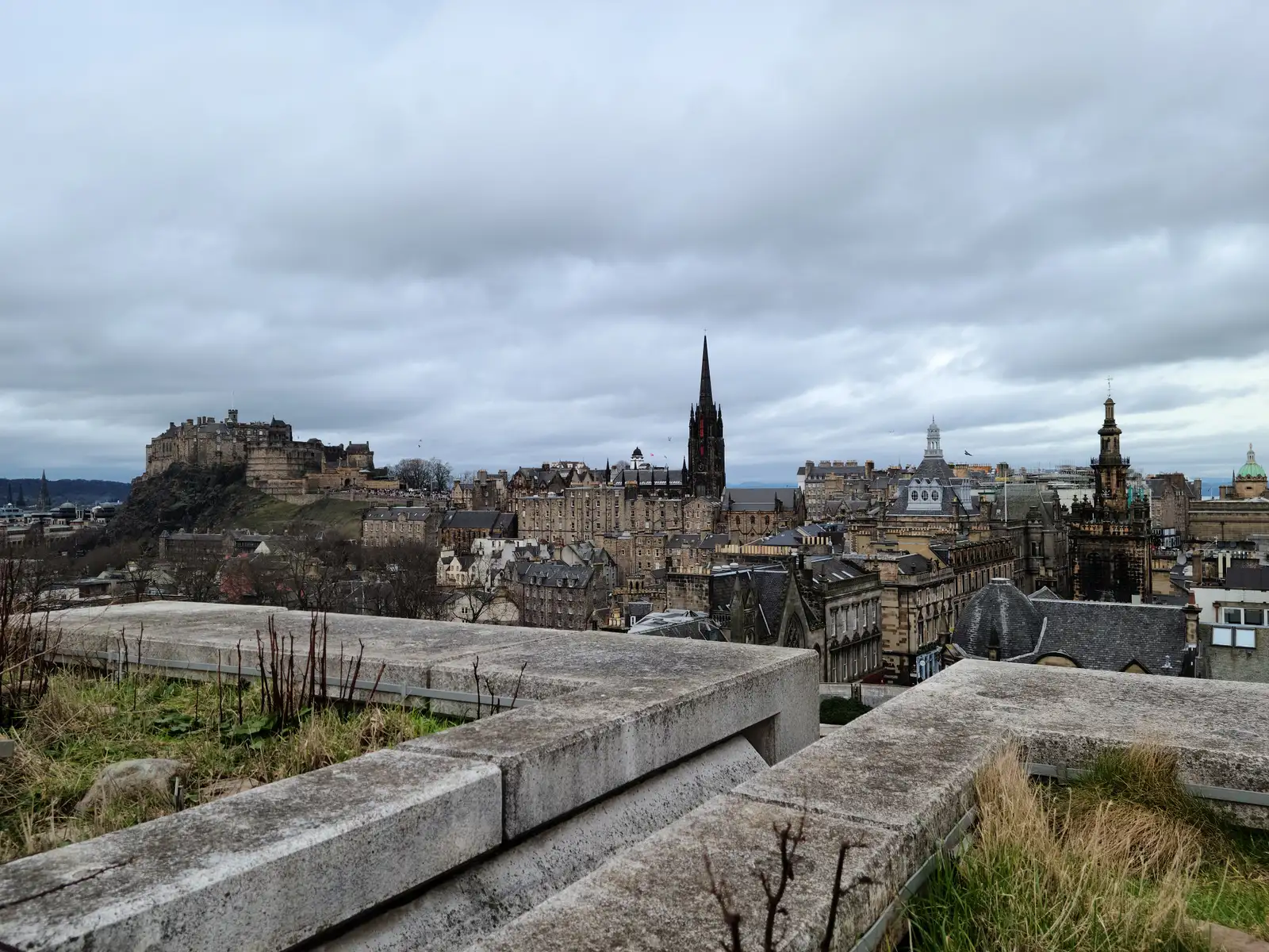 View from Calton Hill across stone parapets to Edinburgh Castle on its crag and the Hub's gothic spire above Old Town rooftops