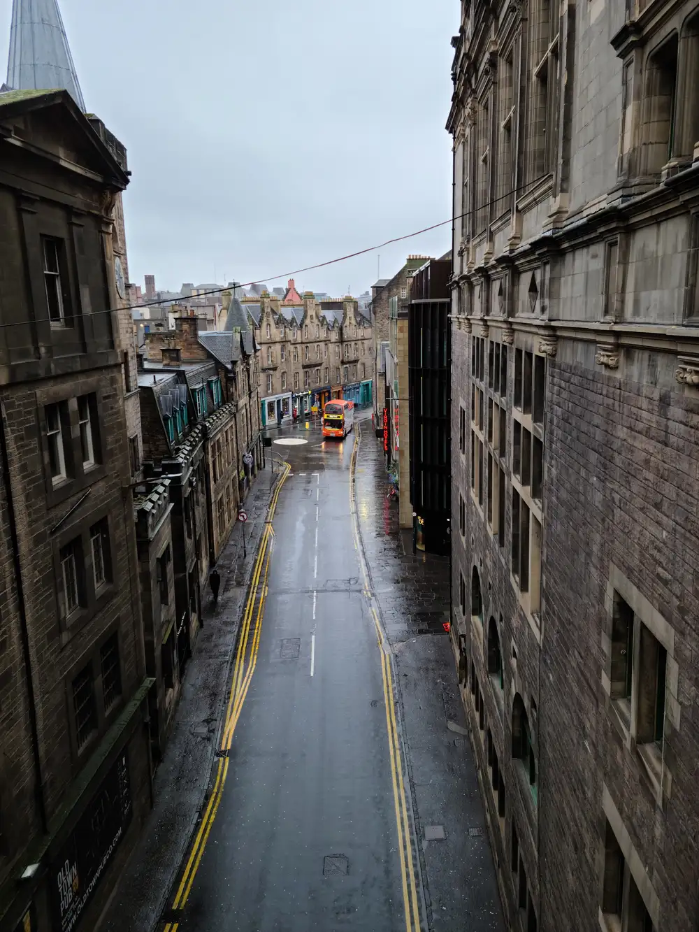 Looking down a rainy Edinburgh Old Town street from above with a red double-decker bus and tenement skyline in the distance