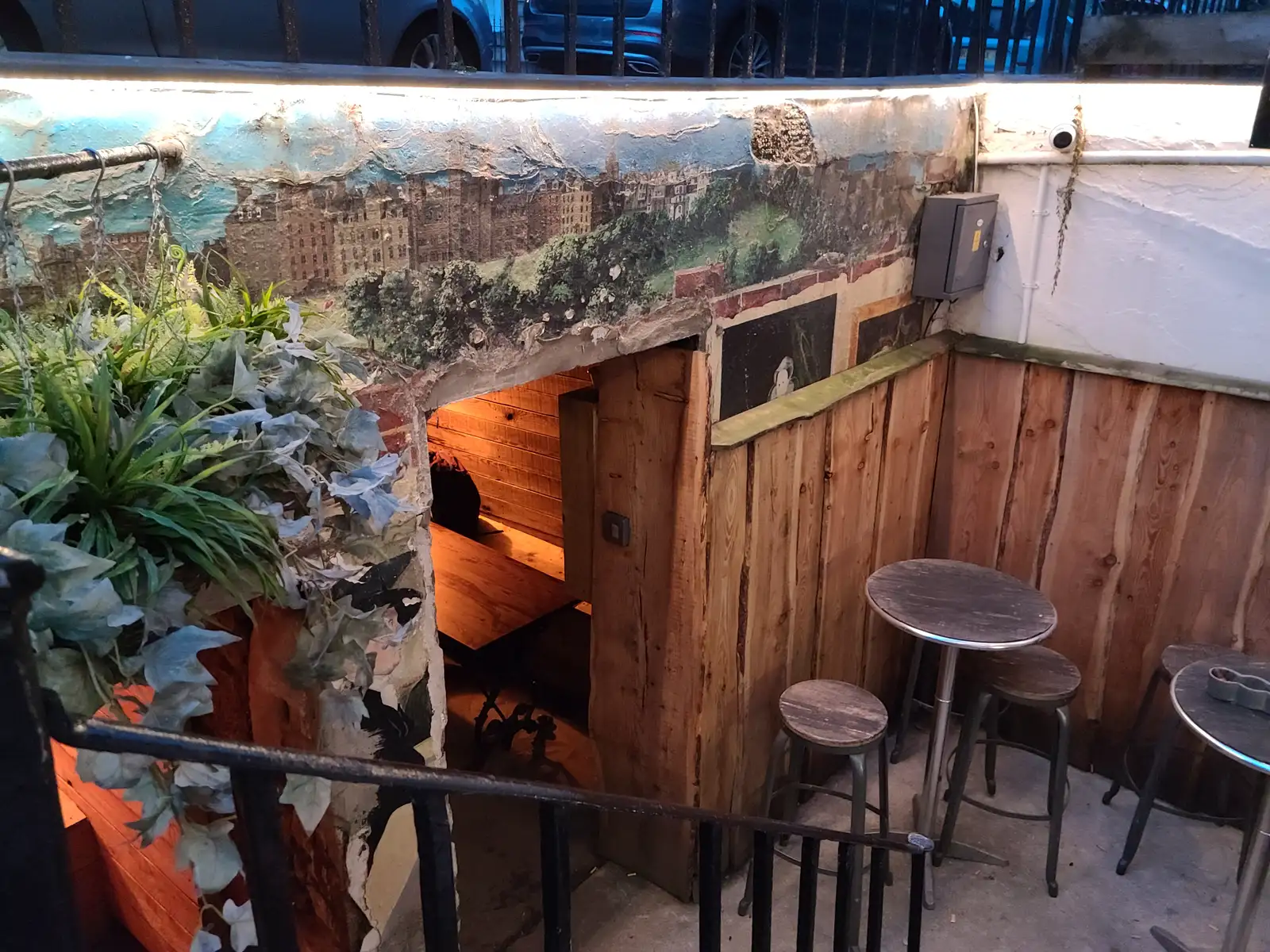 Sunken courtyard bar in Edinburgh Old Town with painted skyline mural, wooden cladding, hanging ivy and metal stools