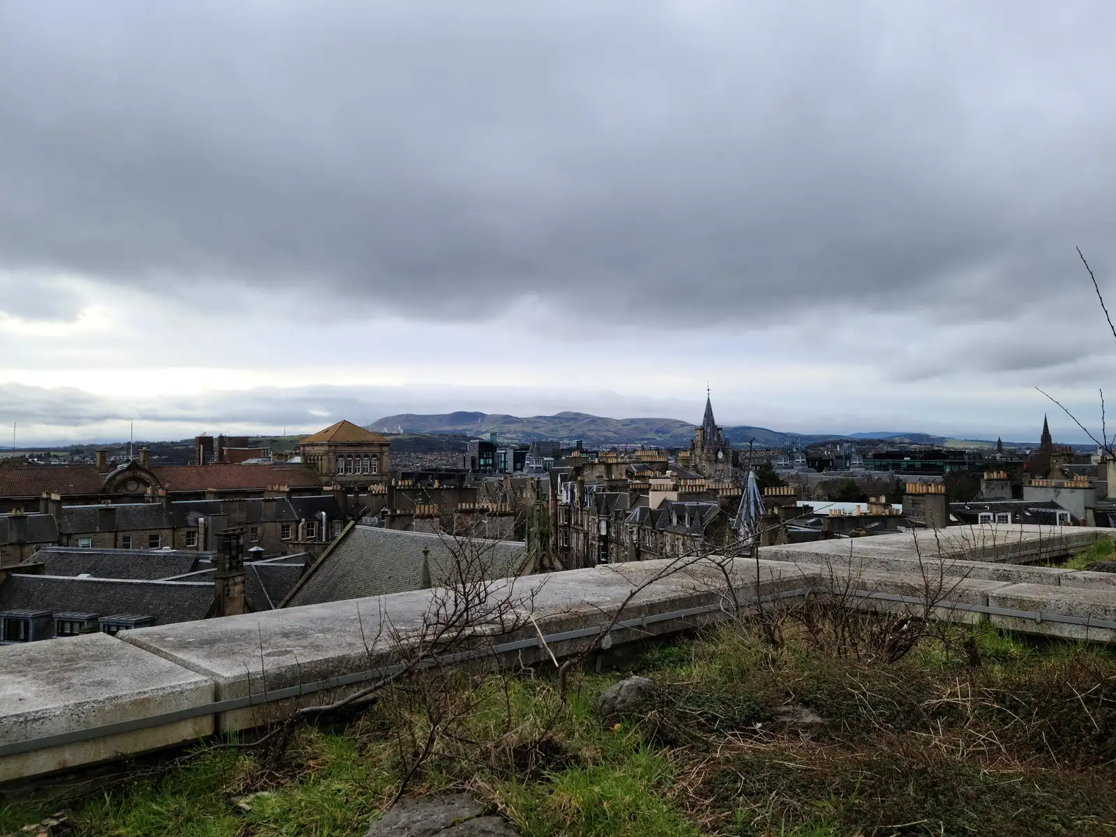 View from Calton Hill across Edinburgh Old Town rooftops and church spires towards the Pentland Hills under gray skies