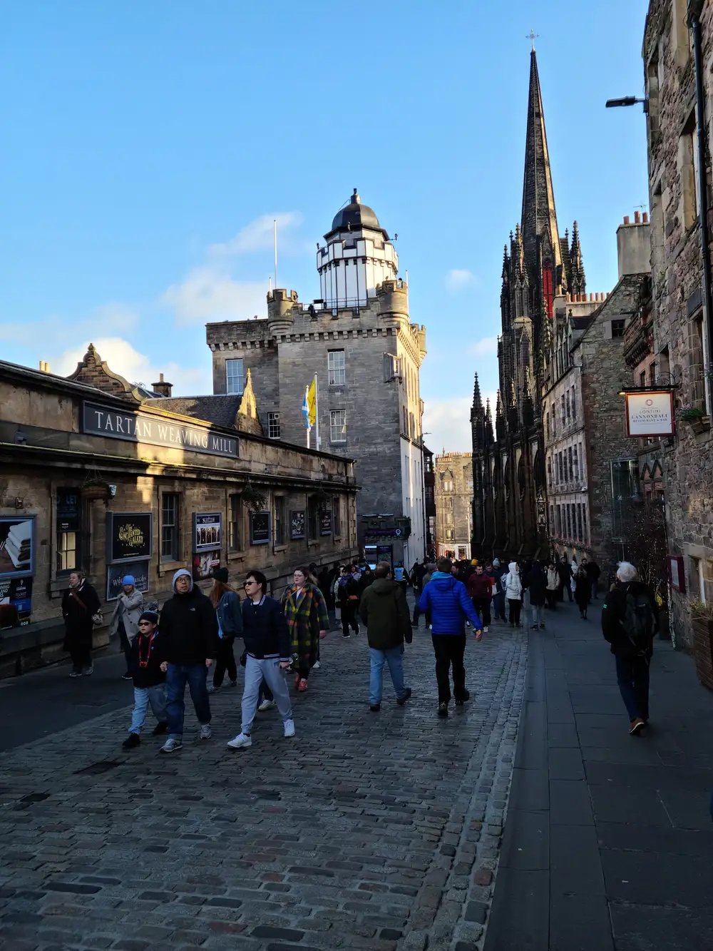 Tourists walking the cobbled Royal Mile in Edinburgh past the Camera Obscura tower and the Hub's gothic spire in winter light