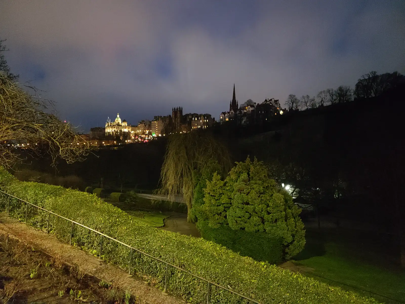 Tolbooth Kirk spire and Old Town silhouette rising above Princes Street Gardens trees under a violet twilight sky in Edinburgh