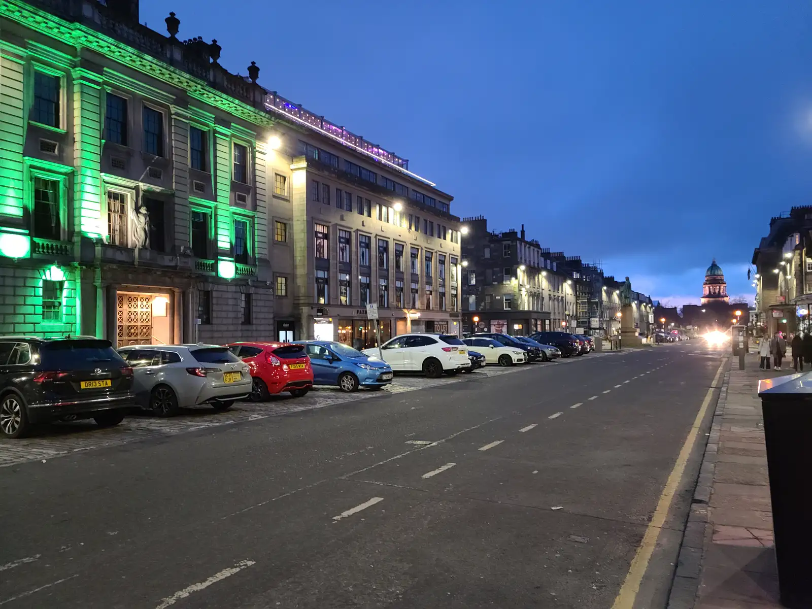 George Street in Edinburgh at dusk with illuminated sandstone facades, parked cars and West Register House dome in the distance