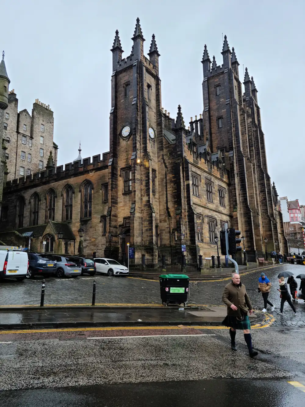 Man in green tartan kilt crosses the rainy street below New College and the Assembly Hall's gothic twin towers on The Mound in Edinburgh