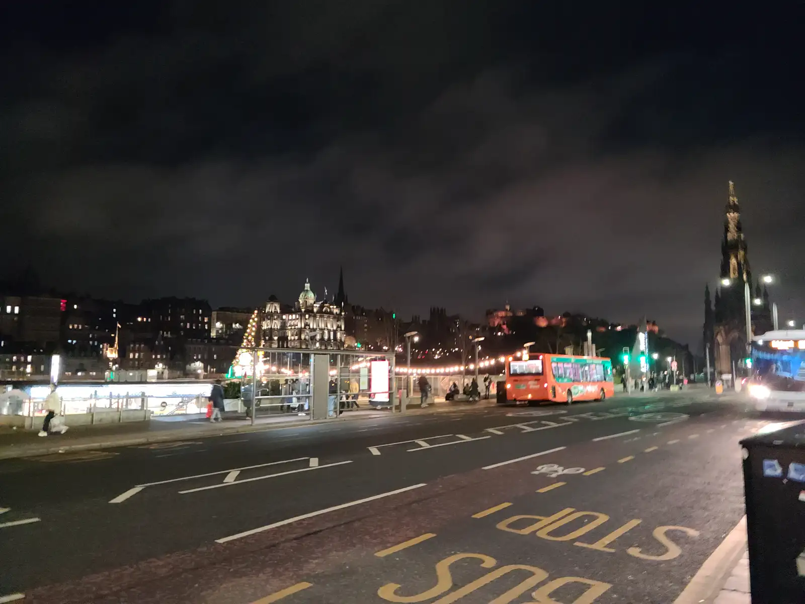 Orange Lothian Buses single-decker at a North Bridge stop in Edinburgh at night with Bank of Scotland dome and Scott Monument lit beyond