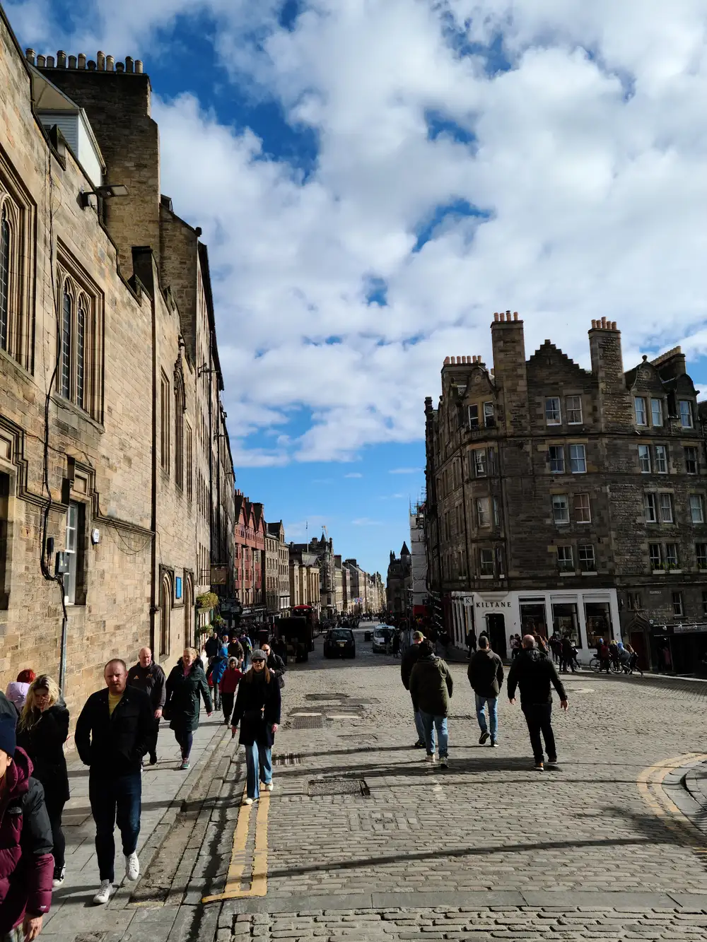 Pedestrians walking the cobbled Royal Mile High Street in Edinburgh past sandstone tenements and Kiltane shop under blue sky