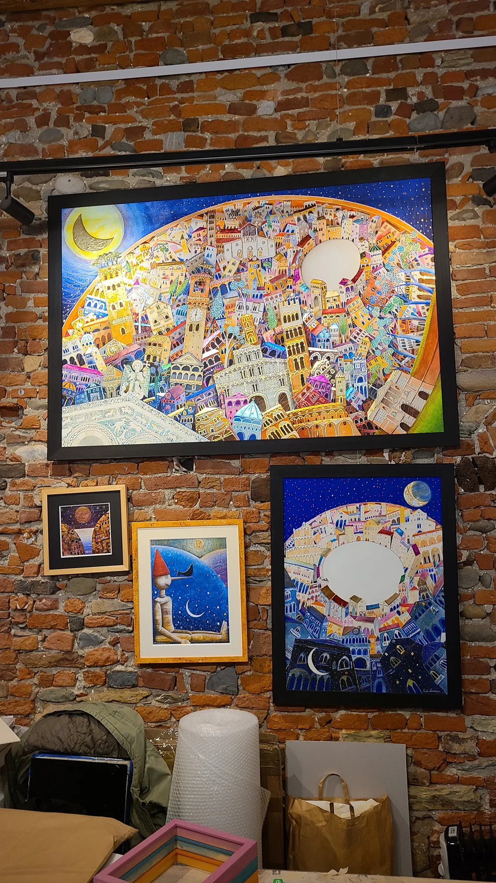 Colorful paintings of a fantastical cityscape with Lucca's towers displayed on a raw brick wall in a local art gallery, vivid blues and golds dominating the canvases
