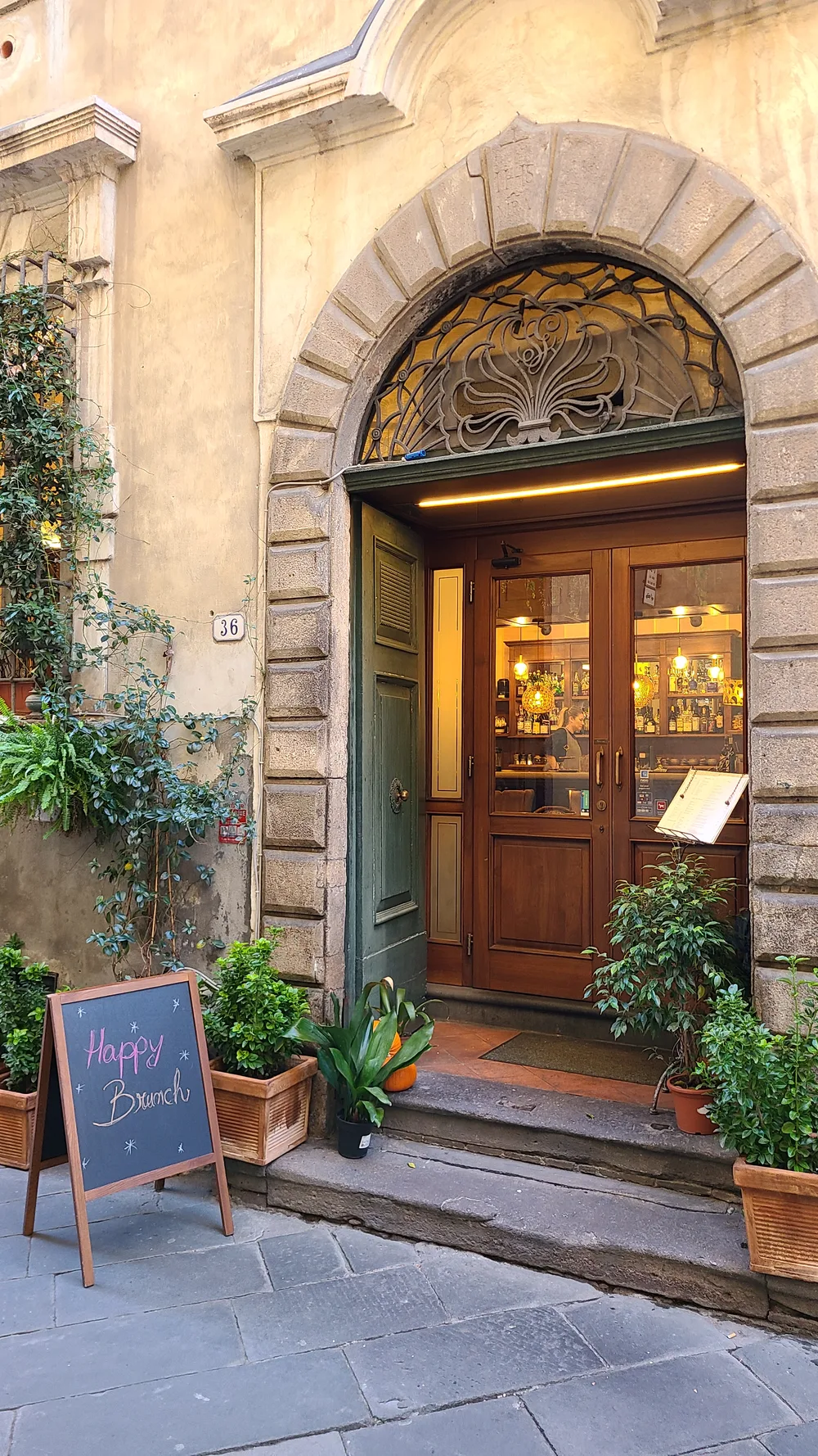 Charming café entrance in Lucca through a stone arch doorway, warm lights glowing inside, potted plants and a Happy Brunch chalkboard sign outside