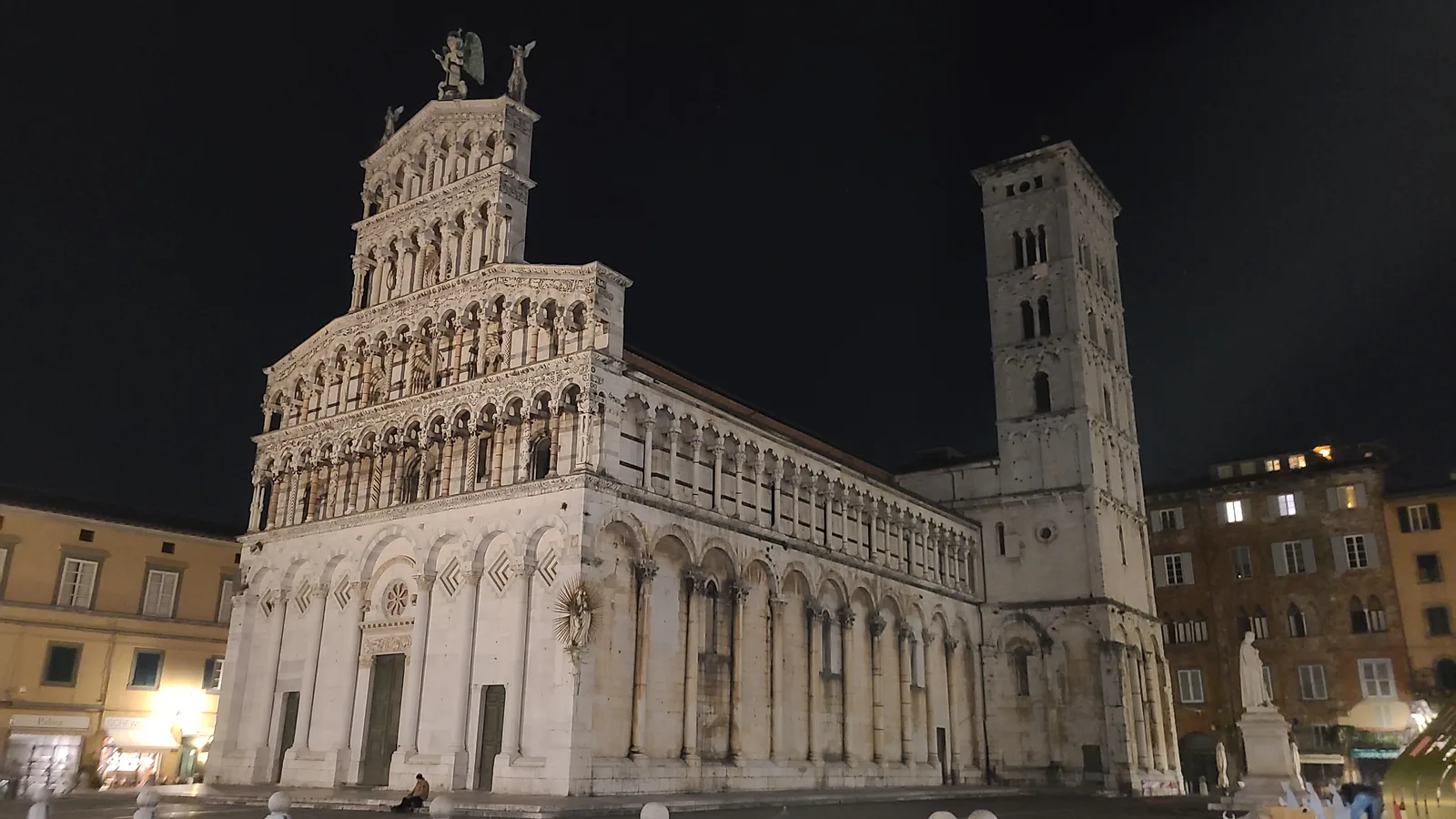 Chiesa di San Michele in Foro floodlit at night in Lucca, the ornate white Romanesque marble facade and bell tower glowing against a black sky, the piazza nearly empty
