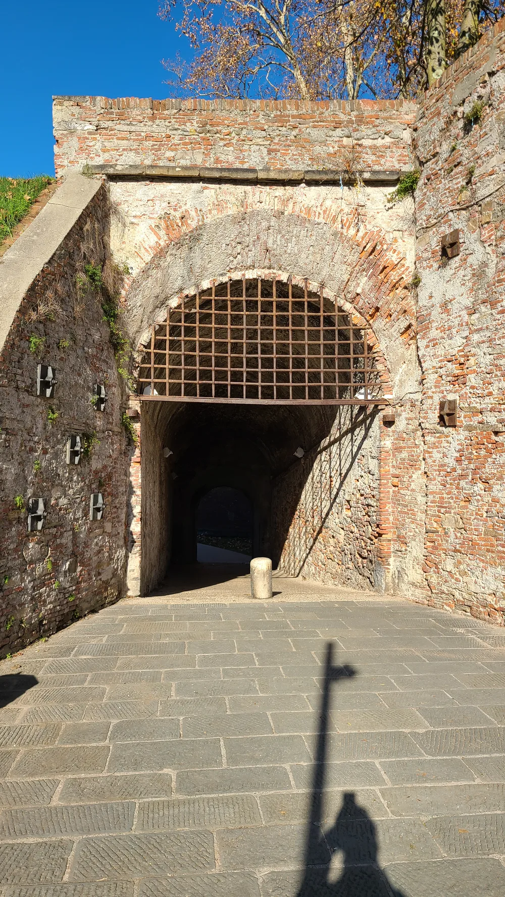 Brick arched passage through Lucca's Renaissance walls with an iron portcullis grating, sunlight visible beyond the dark tunnel entrance