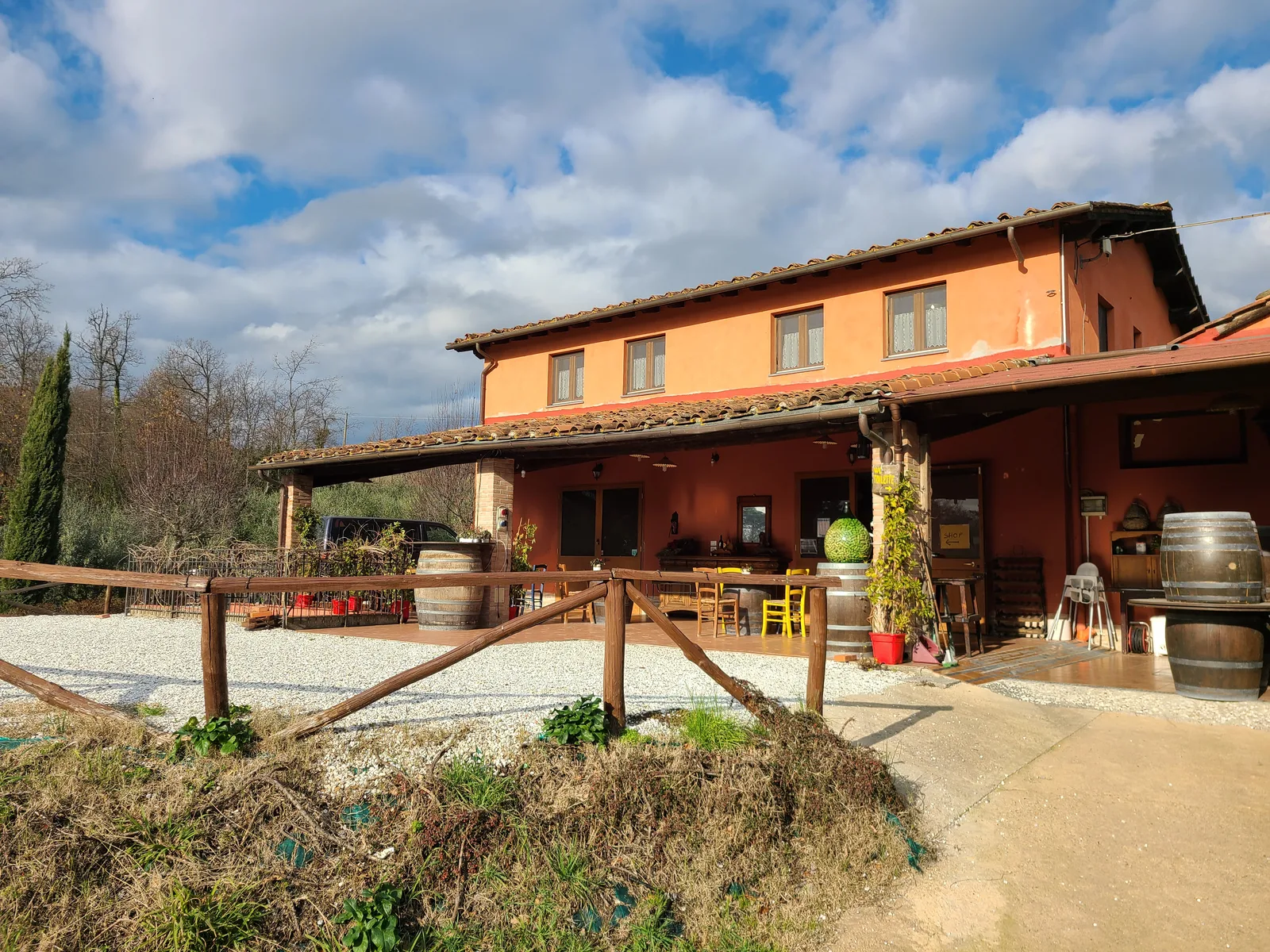 Rustic agriturismo farmhouse exterior at Fattoria al Dotto near Lucca, with wine barrels, outdoor seating area, and cypress trees under a partly cloudy sky