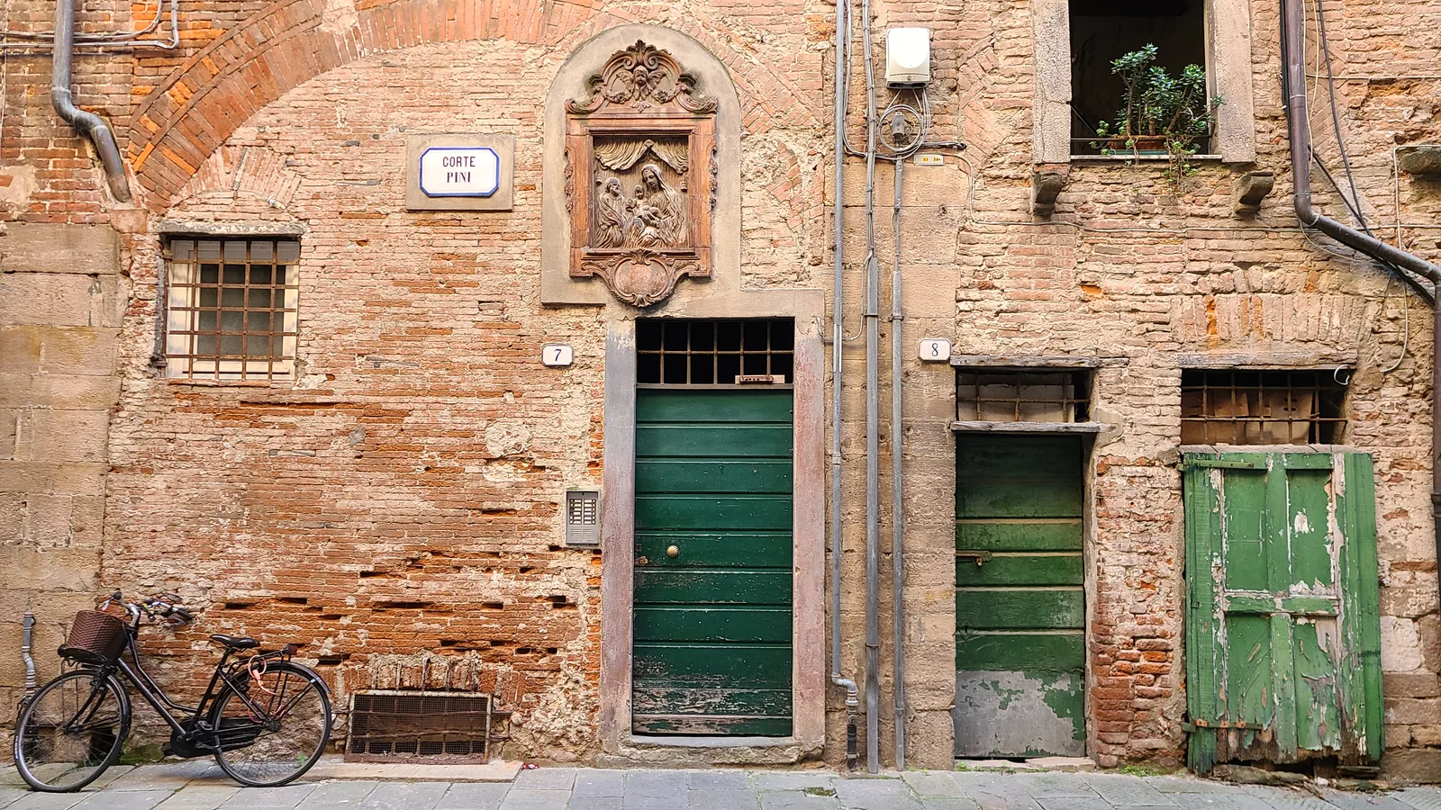 Ancient brick alley in Lucca with two weathered green doors, a bicycle leaning against the wall, and a carved stone relief medallion above the doorway